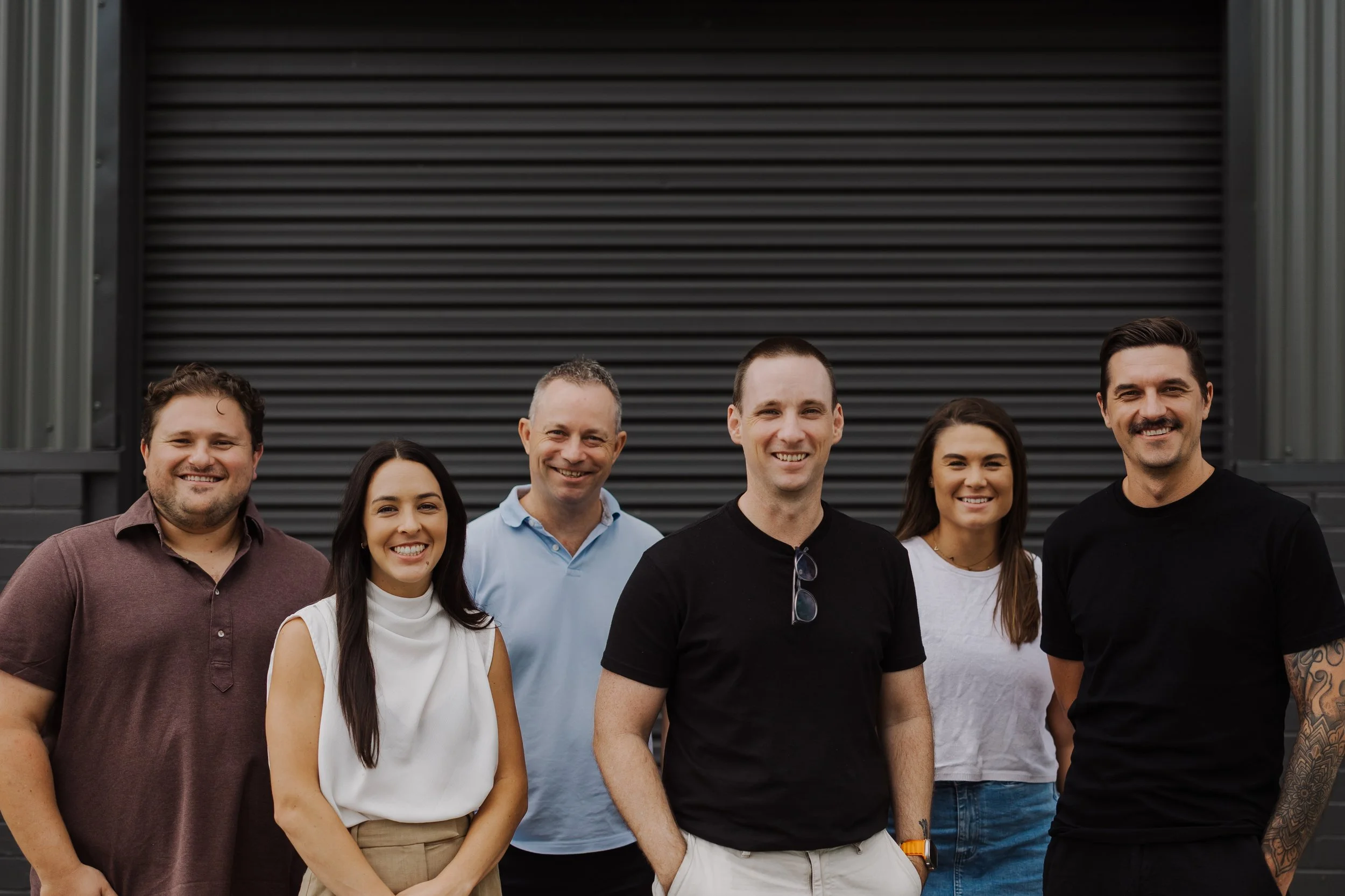 Group of six diverse people standing in front of a dark grey industrial building, smiling at the camera.