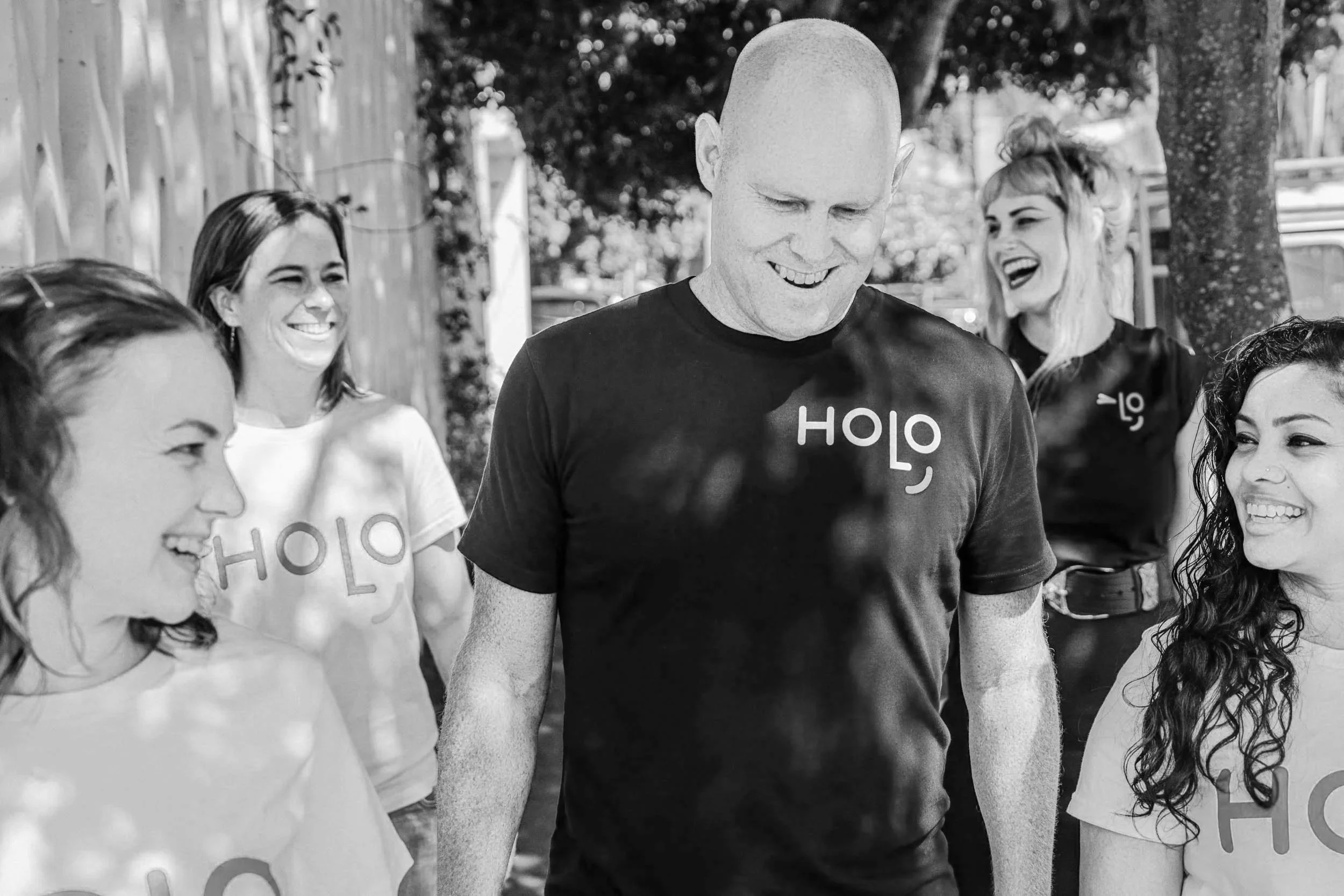 A group of smiling women and a man walking outdoors, some wearing matching T-shirts with the word 'HOLO' on them.