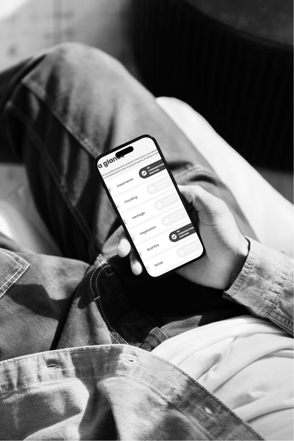 Person sitting with legs crossed, holding a smartphone displaying a property assessment app, black and white photo.