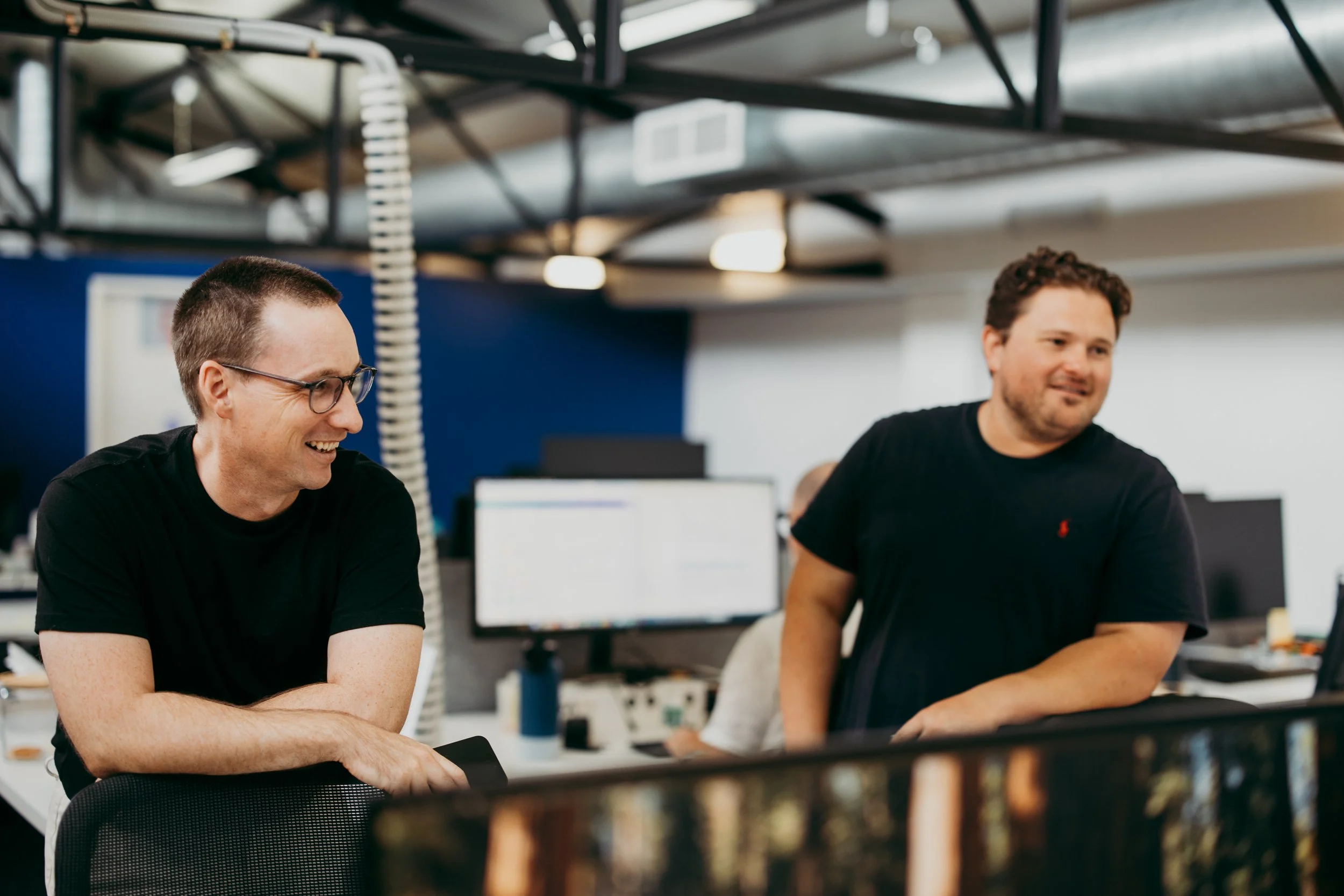 Two men sitting at a table in an office, smiling and engaging in conversation, with computer monitors and office equipment visible in the background.