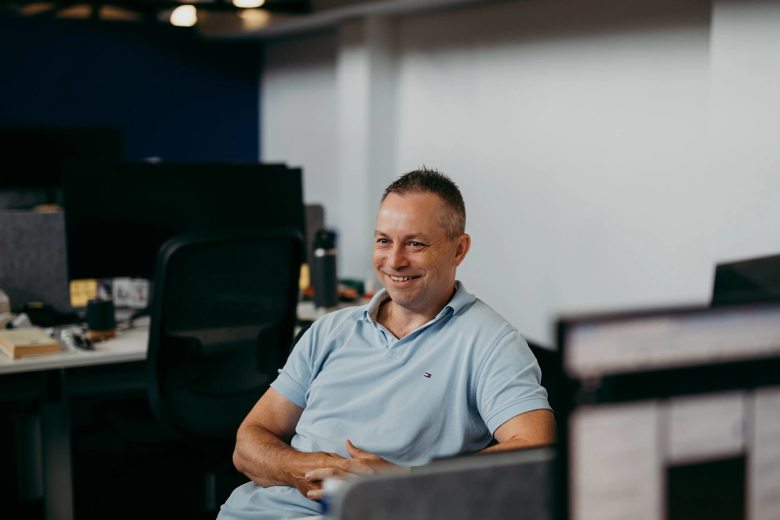 A smiling man sitting in an office environment, wearing a light blue polo shirt, surrounded by computer monitors and office equipment.