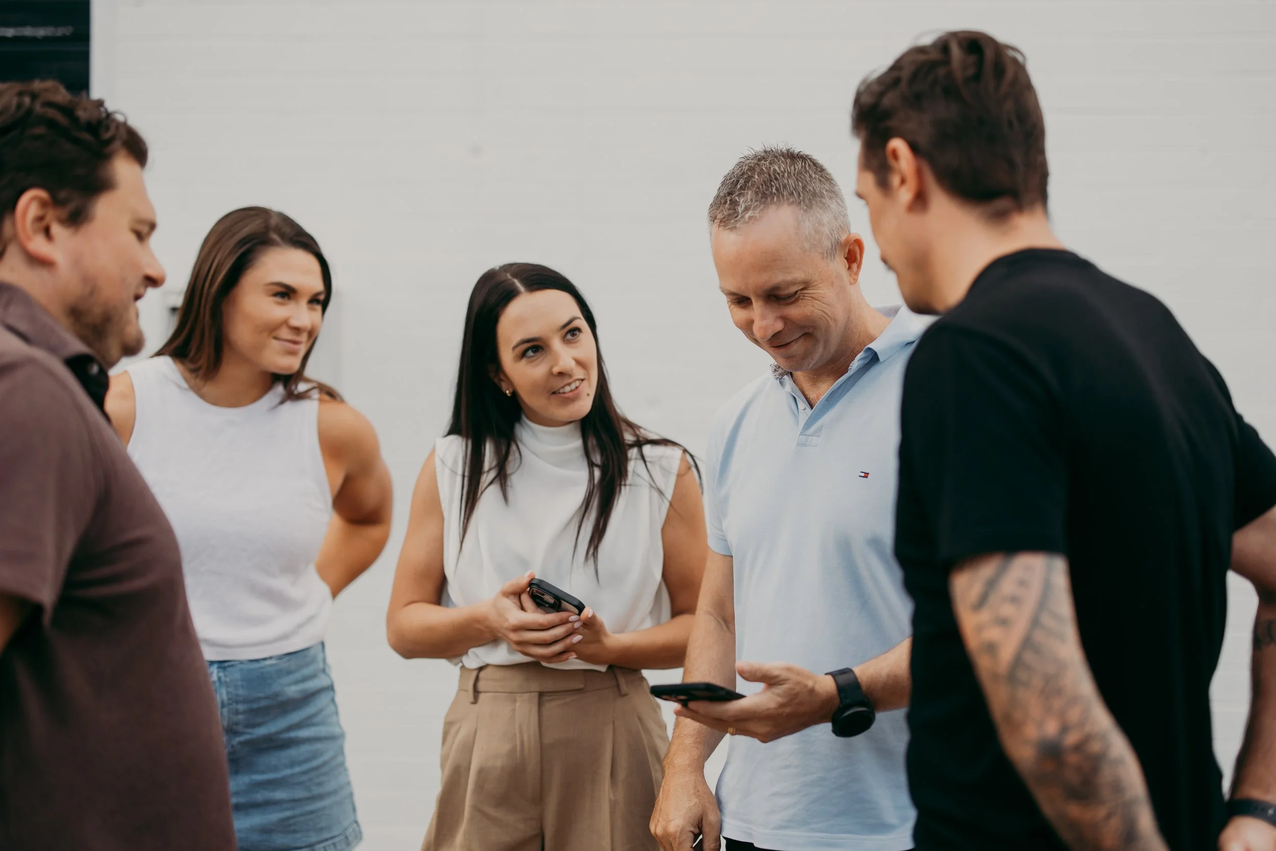 Group of five young adults looking at phone together, standing outdoors in casual clothing, engaged in conversation, with a plain gray wall in the background.
