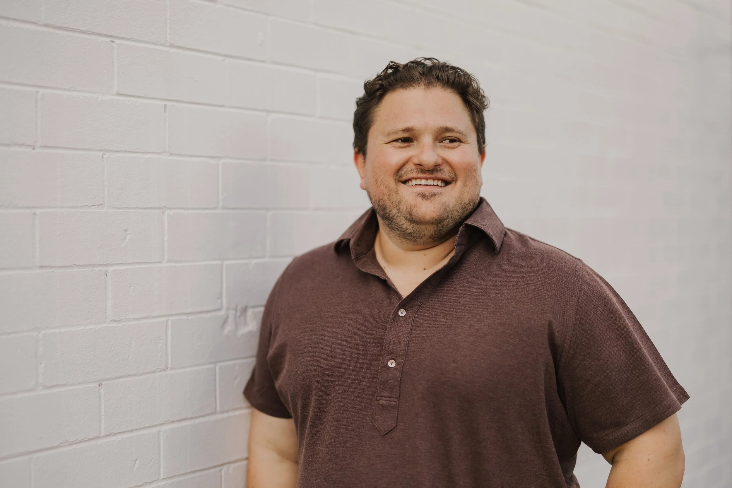 A smiling man with short curly hair and a beard, wearing a maroon button-up shirt, standing against a white brick wall.