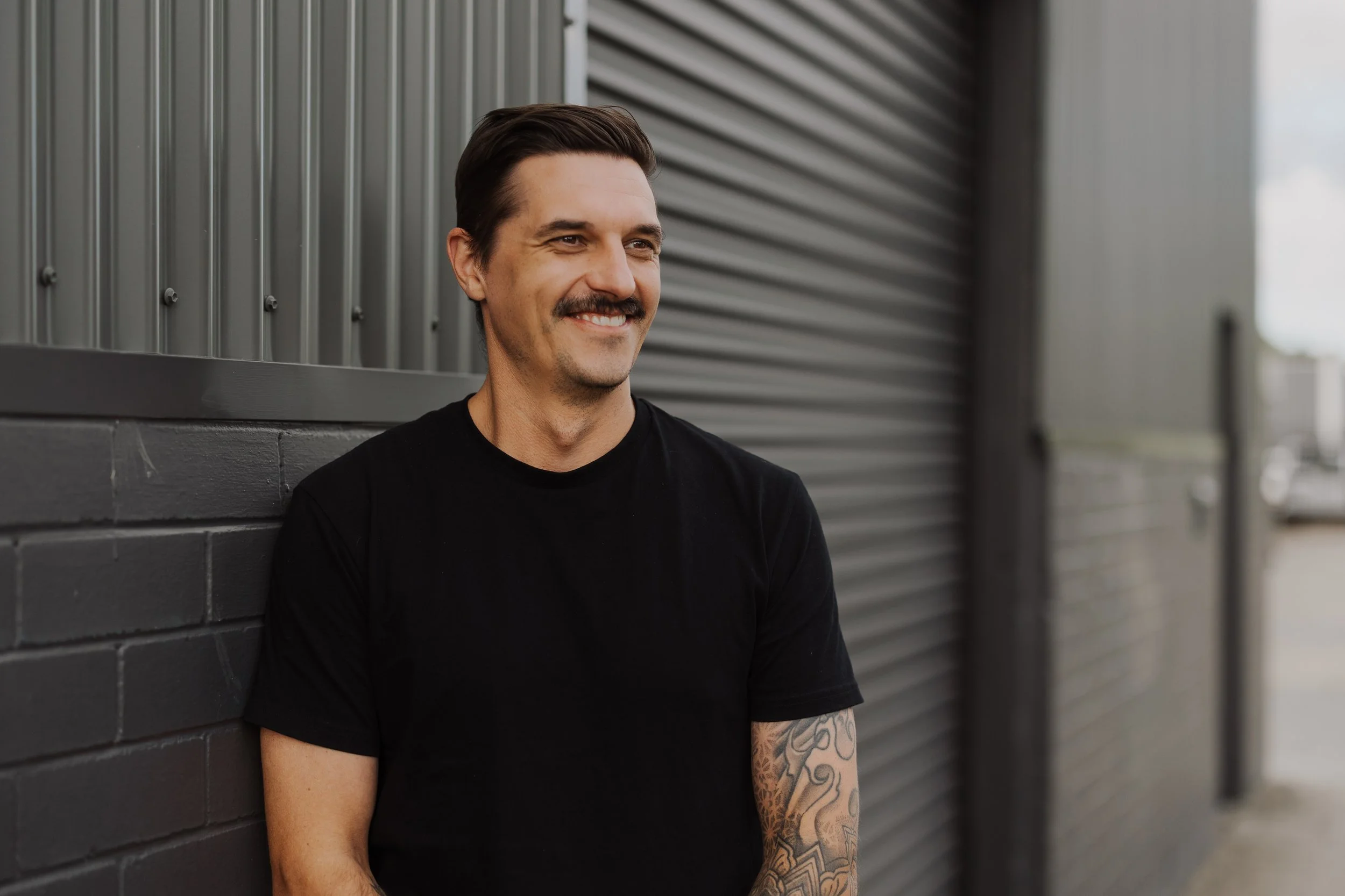 A man with dark hair and a mustache, wearing a black T-shirt, smiling while leaning against a dark gray brick wall with corrugated metal siding in the background.