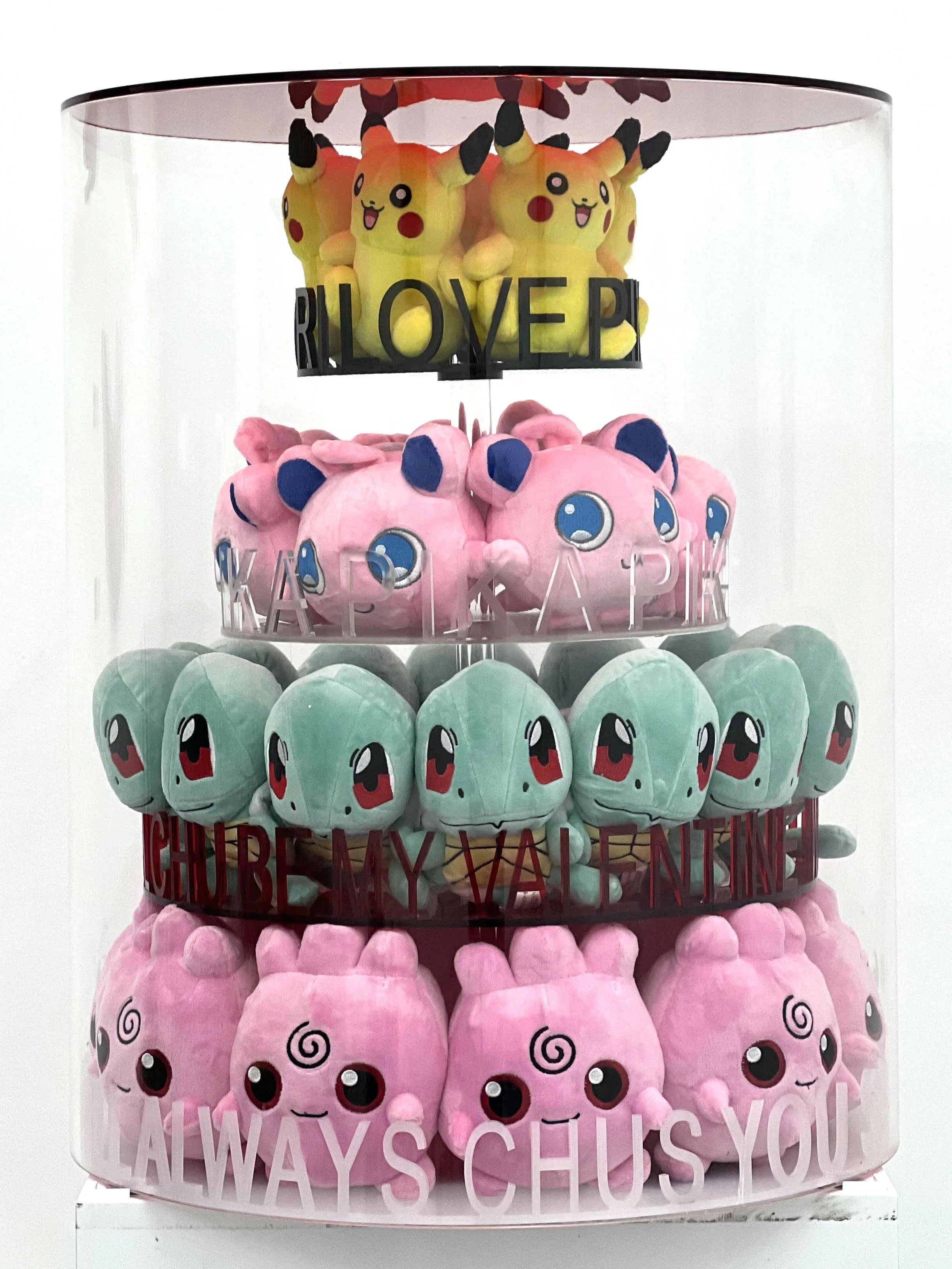 Four tiers of Pokémon plush toys in a glass display case, featuring Pikachu at the top, Mew at the second, Bulbasaur at the third, and Jigglypuff at the bottom, with text on each tier reading, 'I LOVE P,' 'KARDKAPL,' 'CHUBBY VALENTINE,' and 'ALWAYS C