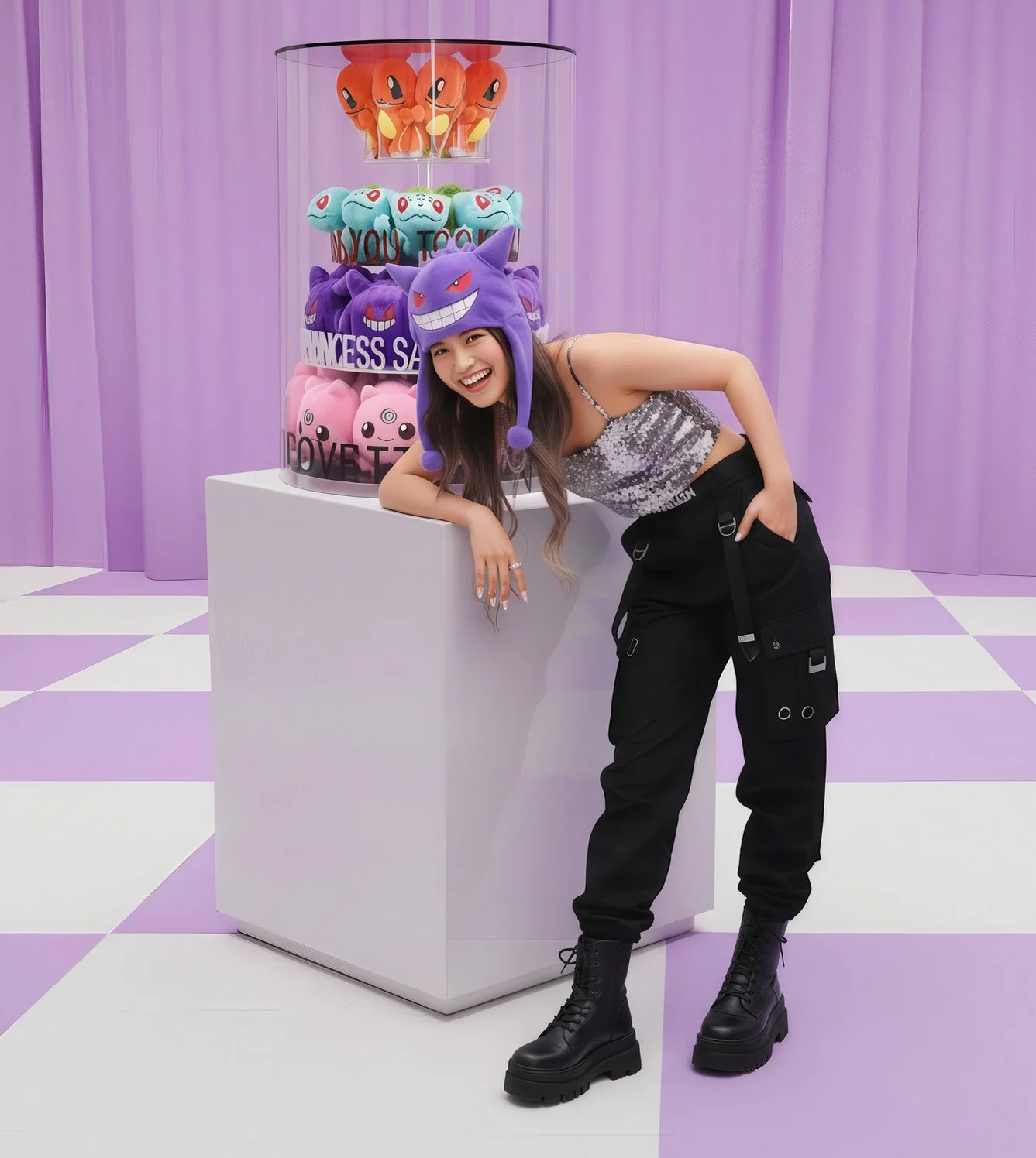 A smiling woman wearing a purple Pokémon Gengar hat, black cargo pants, and black combat boots, leaning on a white pedestal with a display of colorful Pokémon plush toys behind her, set against a purple curtain background.