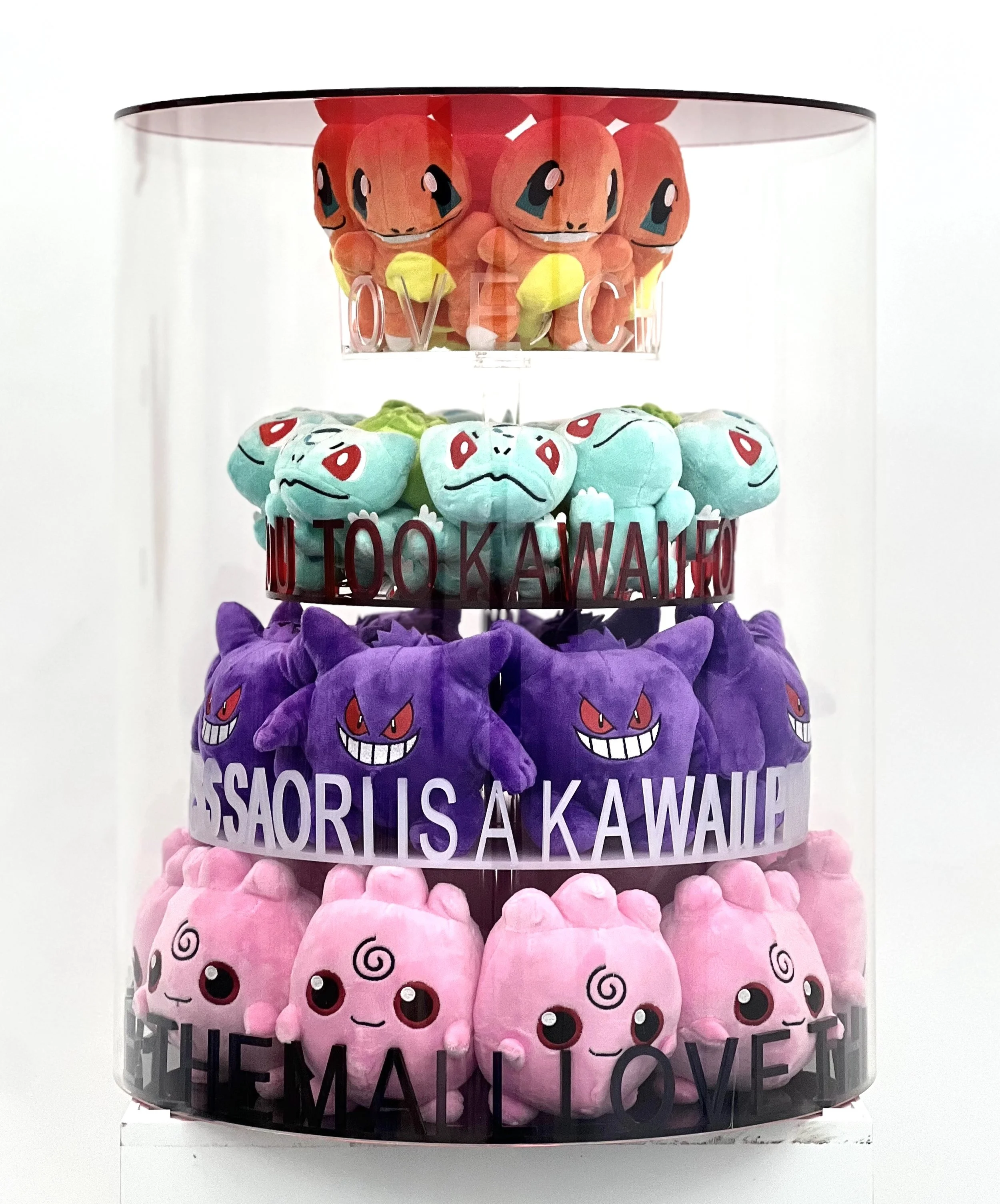 A clear display case with four tiers of plush toys featuring Pokemon characters, from top to bottom: Charmander, Bulbasaur, Gengar, and Jigglypuff, with text on the case including "NOT TOO KAWAII," "GOSSARIS IS A KAWAII," and "THE MALLOVE."