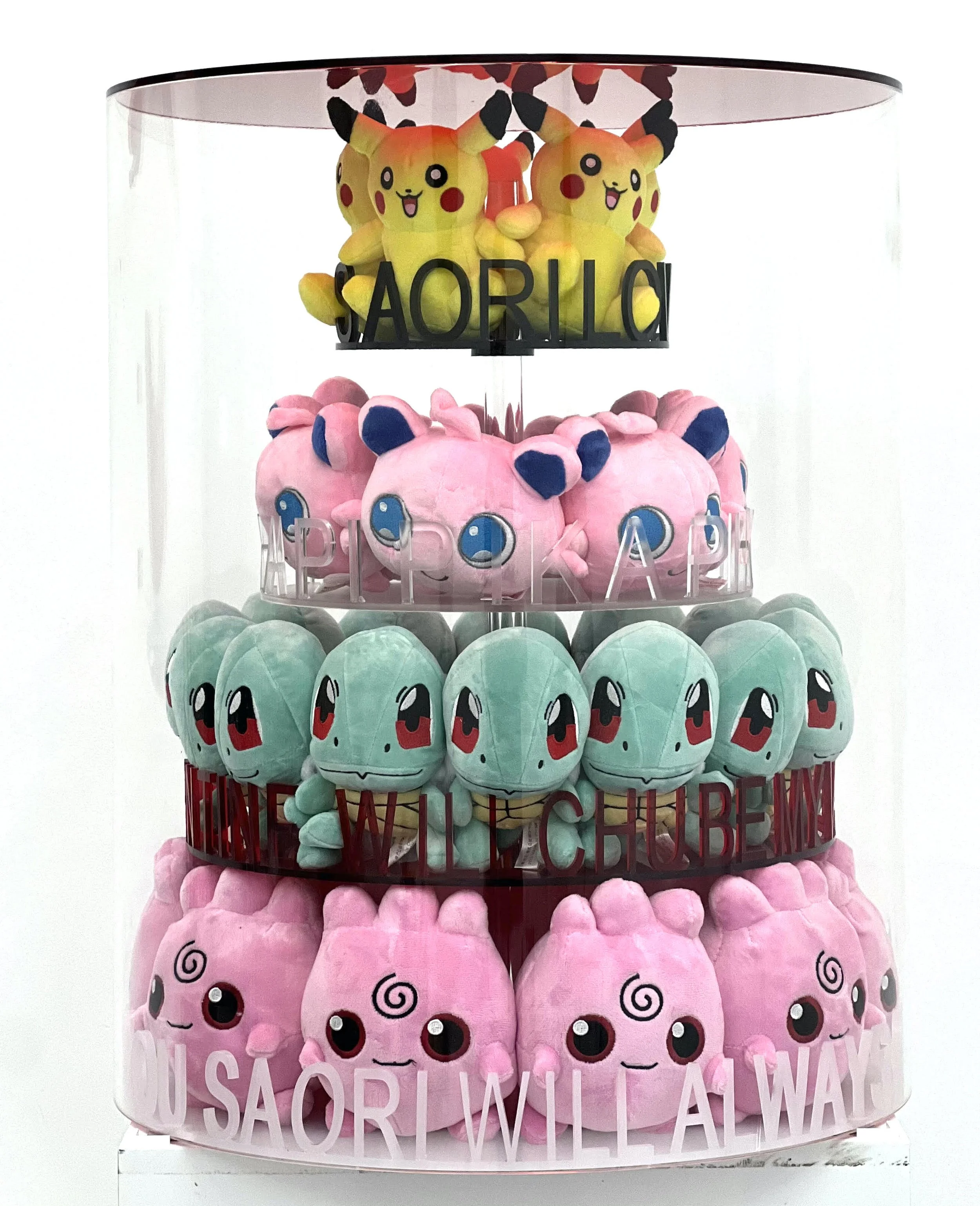 Display of plush toys arranged in four tiers, featuring Pikachu at the top, followed by Jigglypuff, Squirtle, and more plush characters, with slogans like 'SAORU,' 'HAPPINESS,' 'WILL ALWAYS,' and 'YOU SAORI WILL ALWAYS' written on the display.