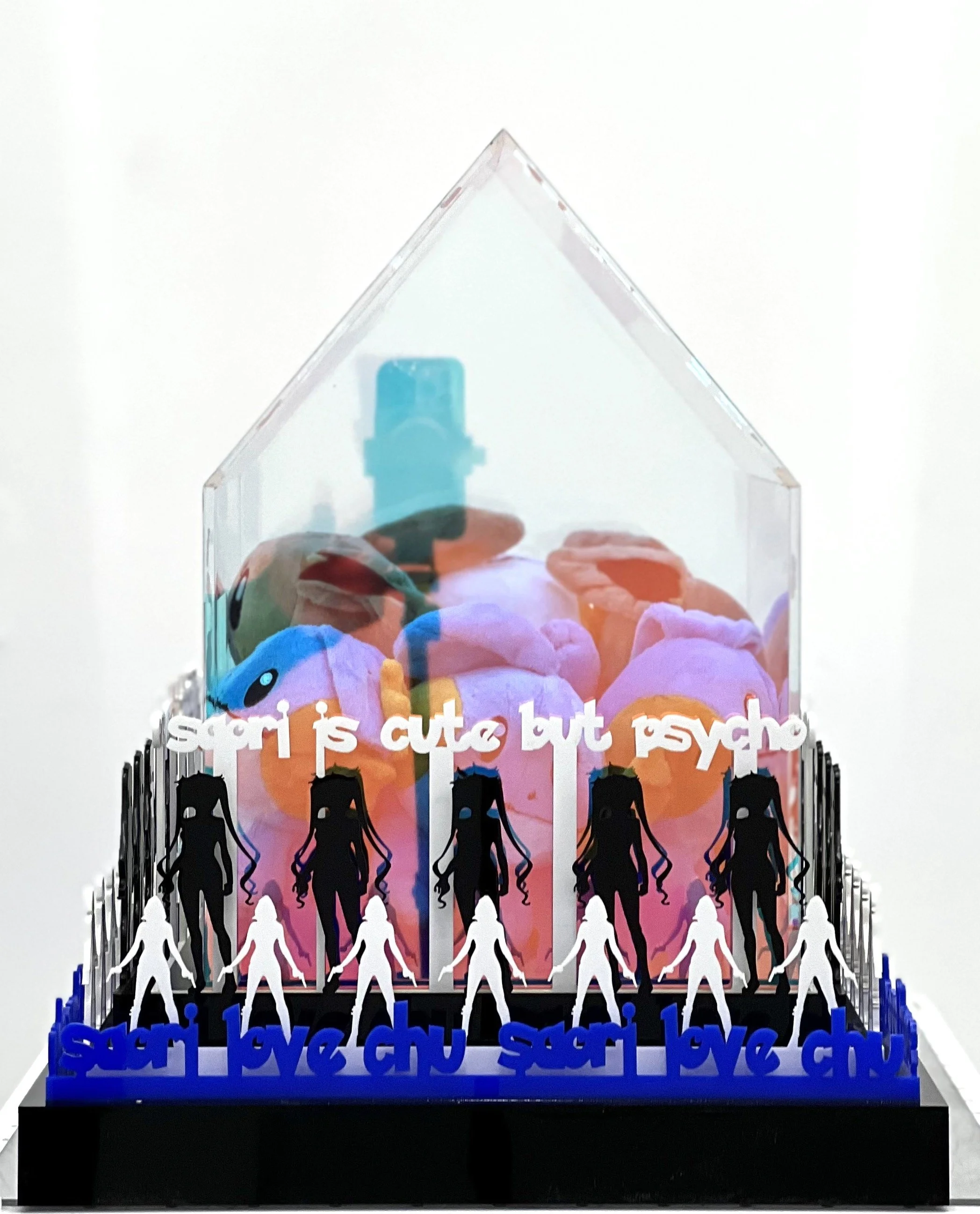 A decorative display featuring a clear house-shaped container filled with colorful plush toys. The base has silhouette figures of women holding hands, and words like 'siori,' 'love,' 'chu,' and 'psycho' are repeated in blue and black text.