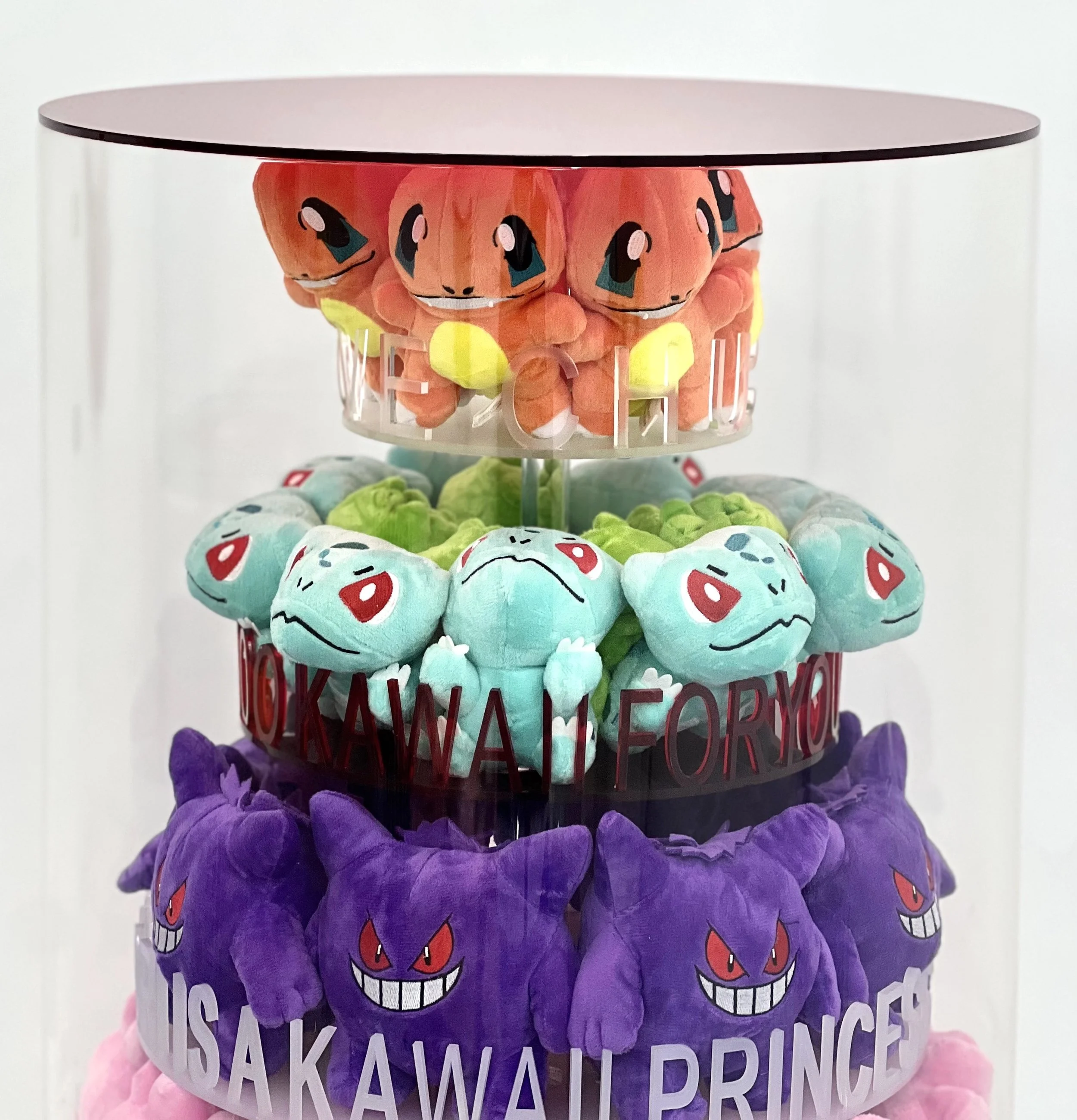 Stack of plush toys representing Pokémon characters, including Charmander, Bulbasaur, Gengar, and Gyarados, arranged on a multi-tiered display with colorful designs and text.