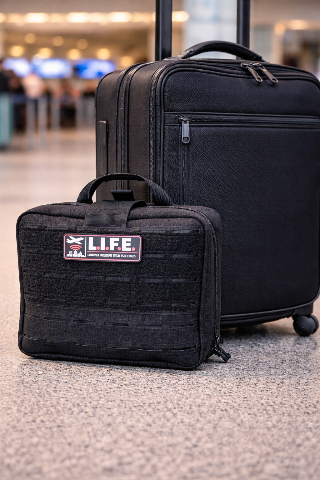A black rolling suitcase and a smaller black travel bag with an 'L.I.F.E' patch, sitting on airport carpet in busy terminal.