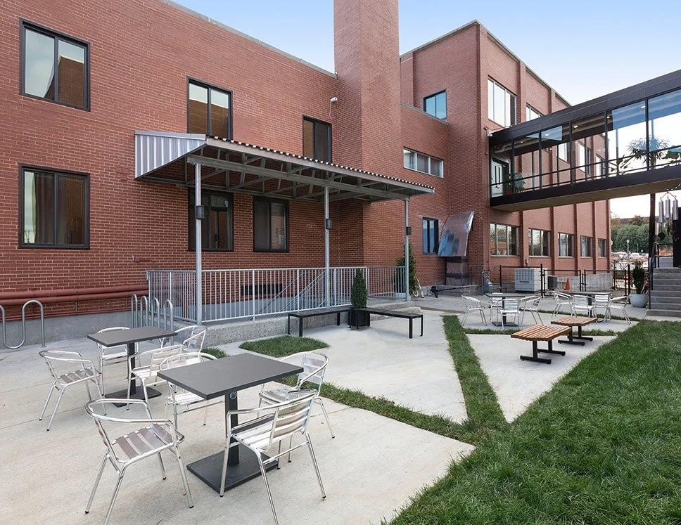 Outdoor patio with tables, chairs, benches, and potted plants next to a brick building with a glass walkway bridge.