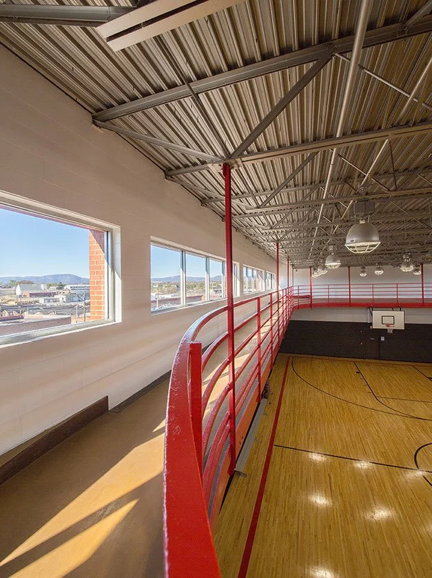 Indoor basketball gym with a polished wooden court, red safety railing on the upper level, large windows on the left, and ceiling lights.