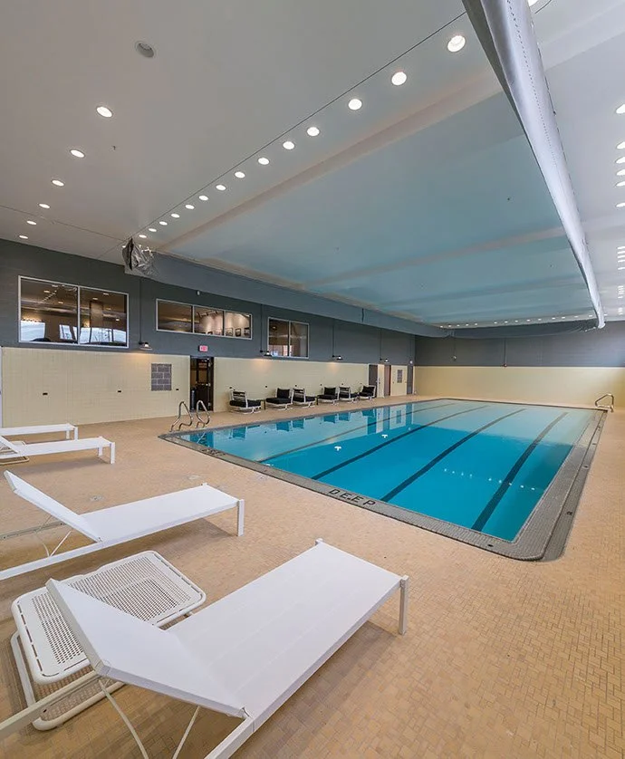 Indoor swimming pool with lounge chairs and seating area along the wall, doors, and windows above.
