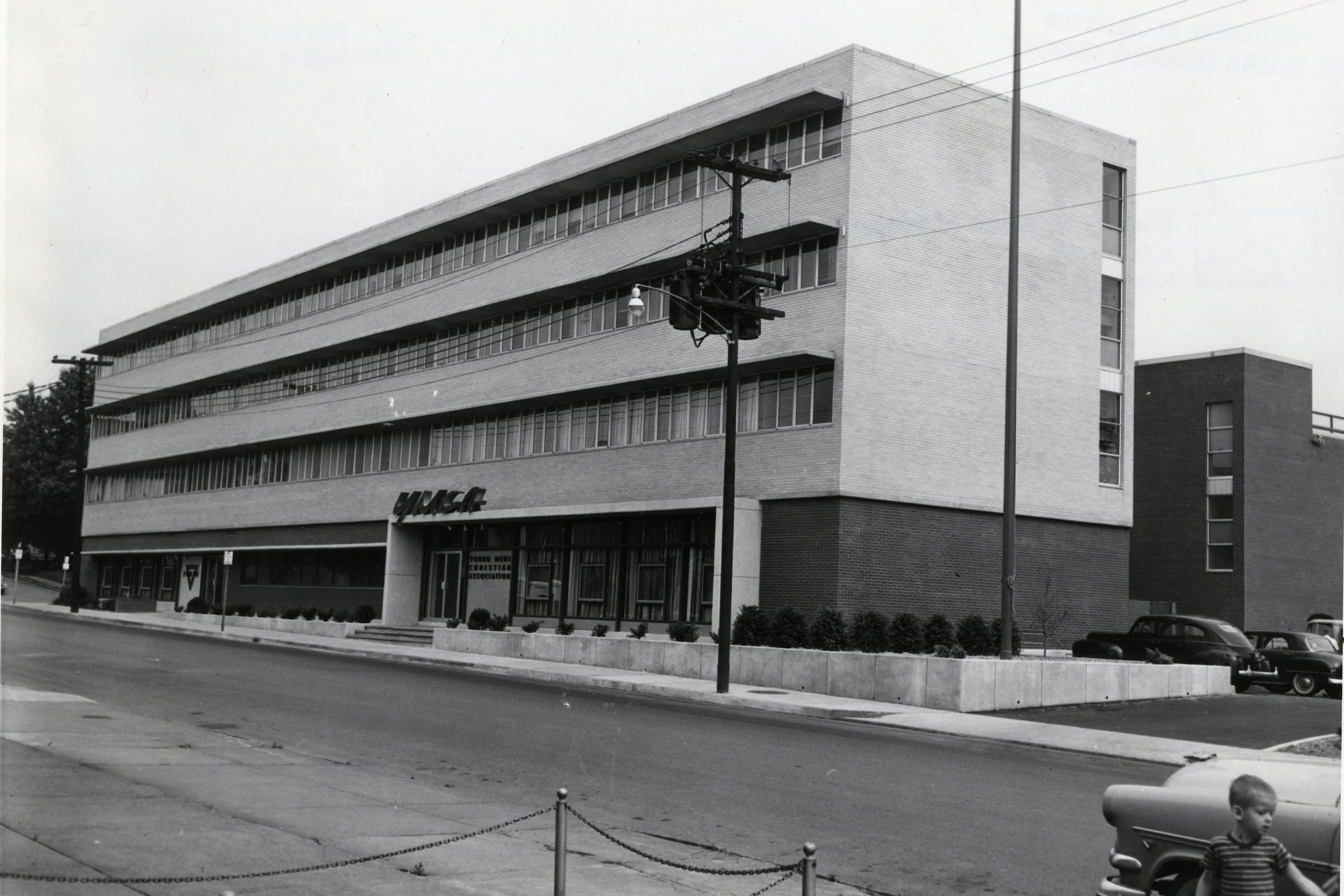 Black and white photo of a modernist brick building with large horizontal windows, identified as the YMCA, with a street and parked cars in the foreground, and a utility pole with power lines.
