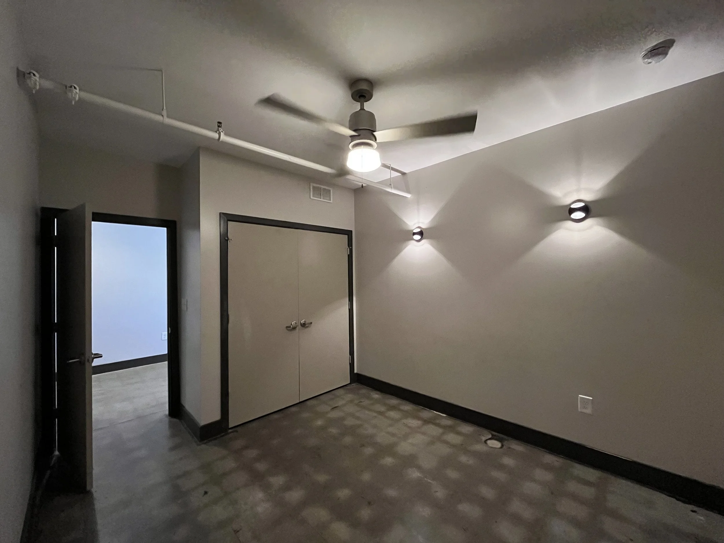Interior of a modern building lobby with a ceiling fan, ceiling lights, a door to the left, and wall-mounted lights casting shadows.