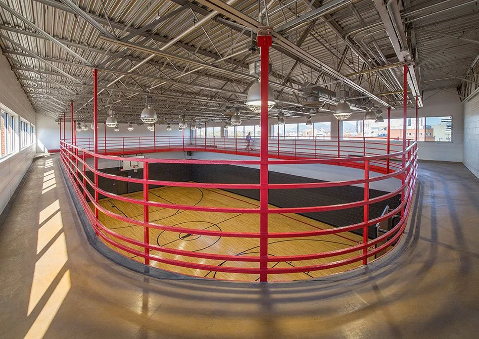 An indoor basketball court with a red railing surrounding it, located in a large, well-lit gymnasium with a high ceiling and multiple windows.