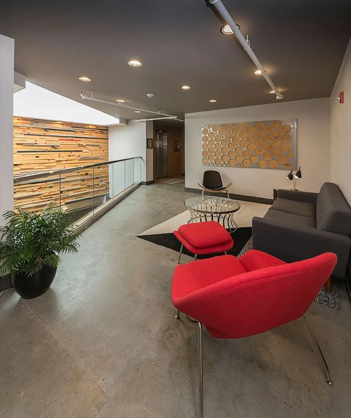 Modern indoor lounge with red armchair, black sofa, glass coffee table, black chair, wall art, potted plant, and elevator in background.