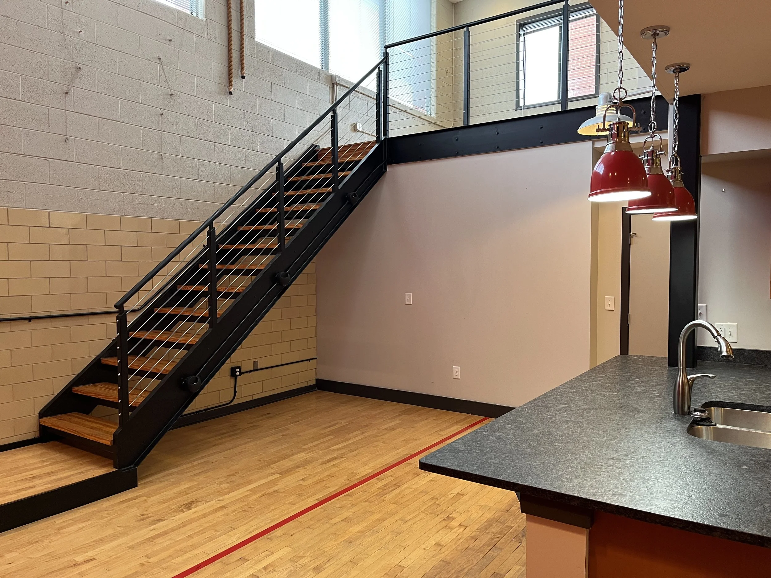 Interior view of a modern kitchen and staircase with black metal railings and wooden steps, featuring red pendant lights hanging above a countertop.