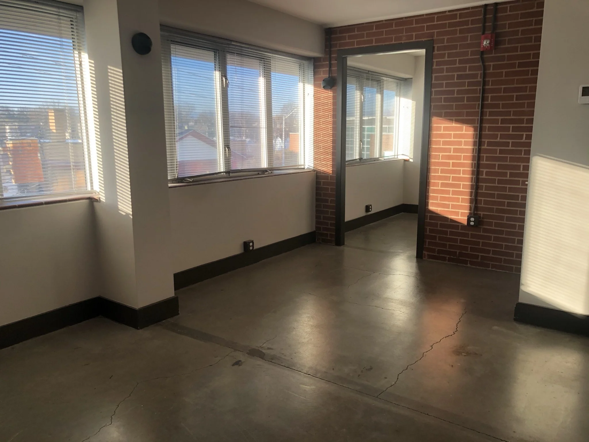Interior of an apartment or office with large windows, brick accent wall, concrete floor, and some electrical outlets.