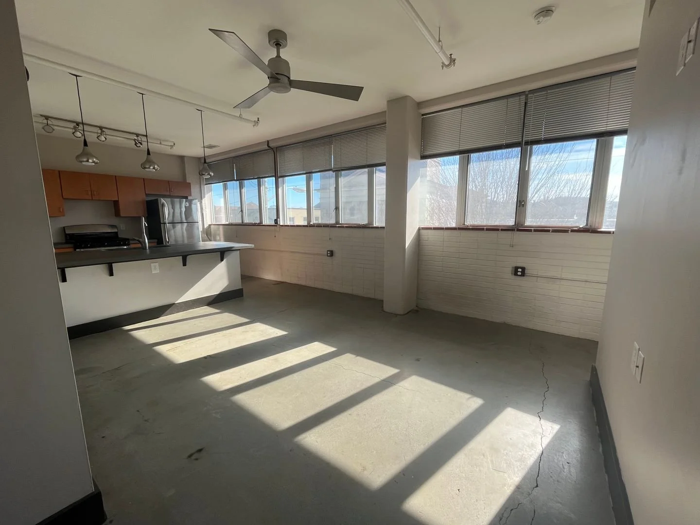 Empty room with large windows, a small kitchen area with a gray counter, wooden cabinets, stainless steel refrigerator, stove, hanging pendant lights, ceiling fan, and exposed concrete floor with cracks.