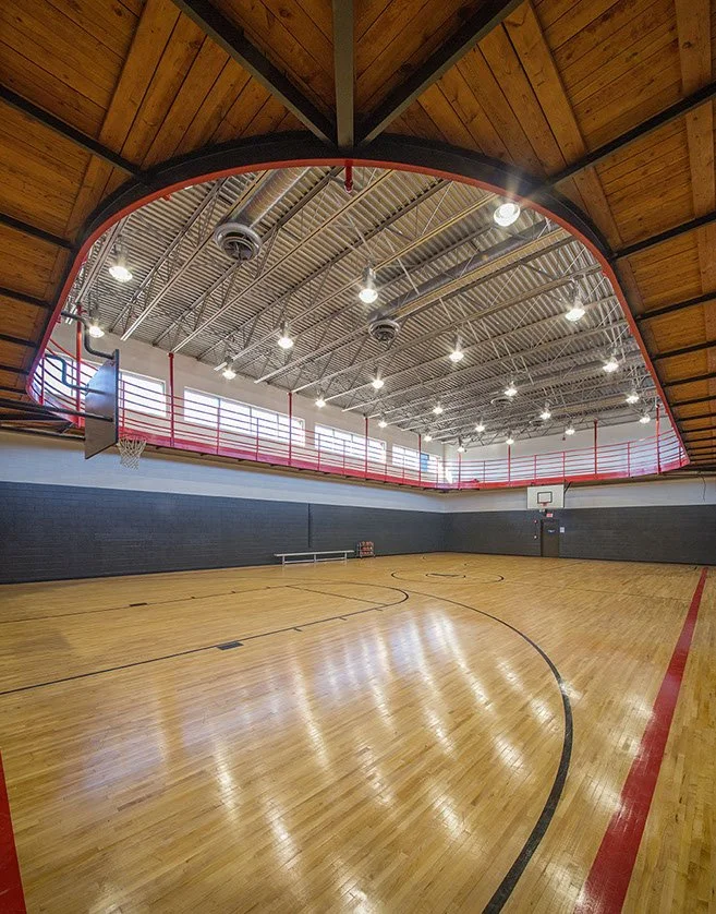 Indoor basketball court with wooden flooring, gray brick walls, basketball hoops, and a red safety railing around the mezzanine area with large windows letting in natural light.
