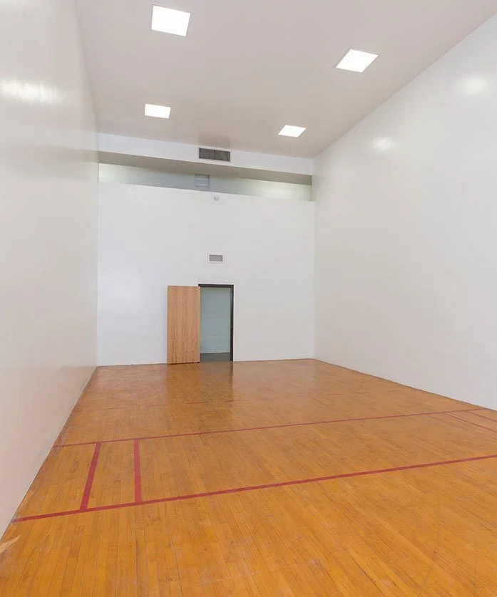 An empty indoor racquetball court with white walls, a wooden floor, and ceiling lights.