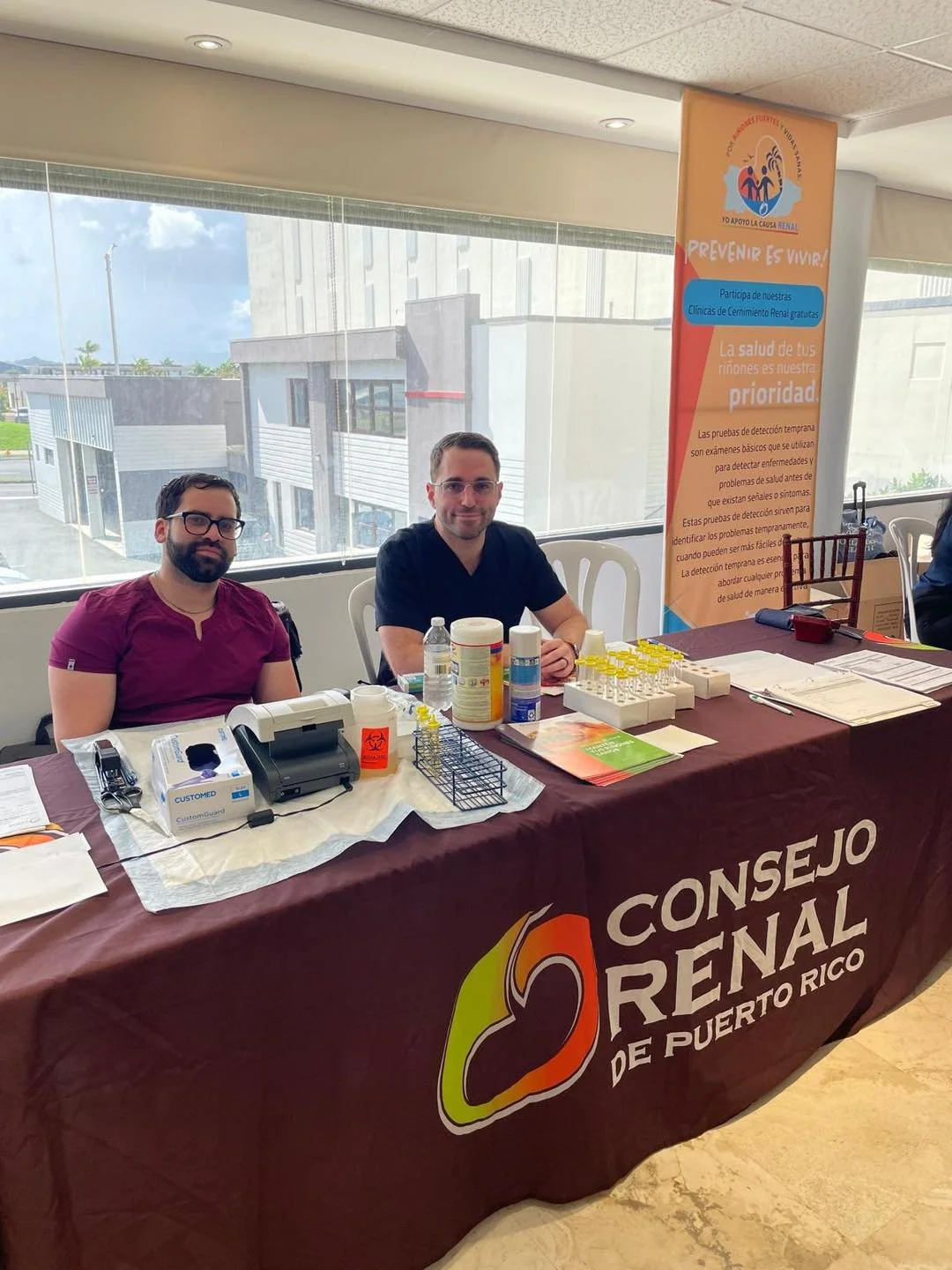 Two men sitting at a table with medical supplies, healthcare posters, and documents, representing a health awareness event by Consejo Renal de Puerto Rico.