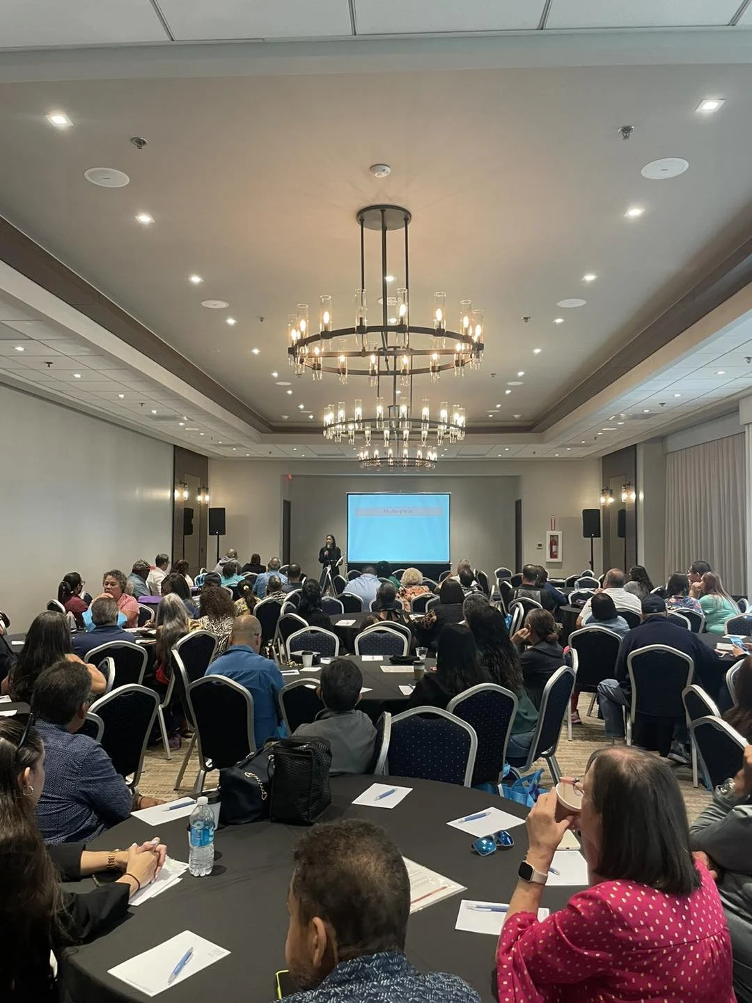 A conference room filled with attendees sitting at round tables, watching a speaker on stage with a presentation screen behind them.