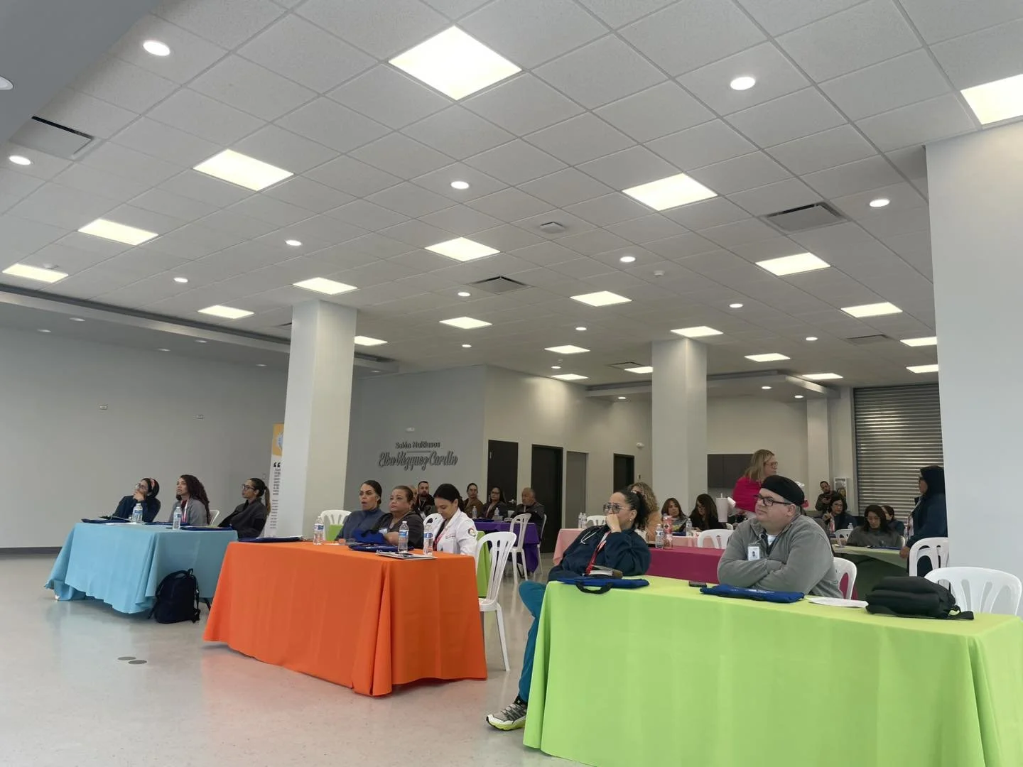 People attending a conference or seminar seated at tables with colorful tablecloths in a large, well-lit room.