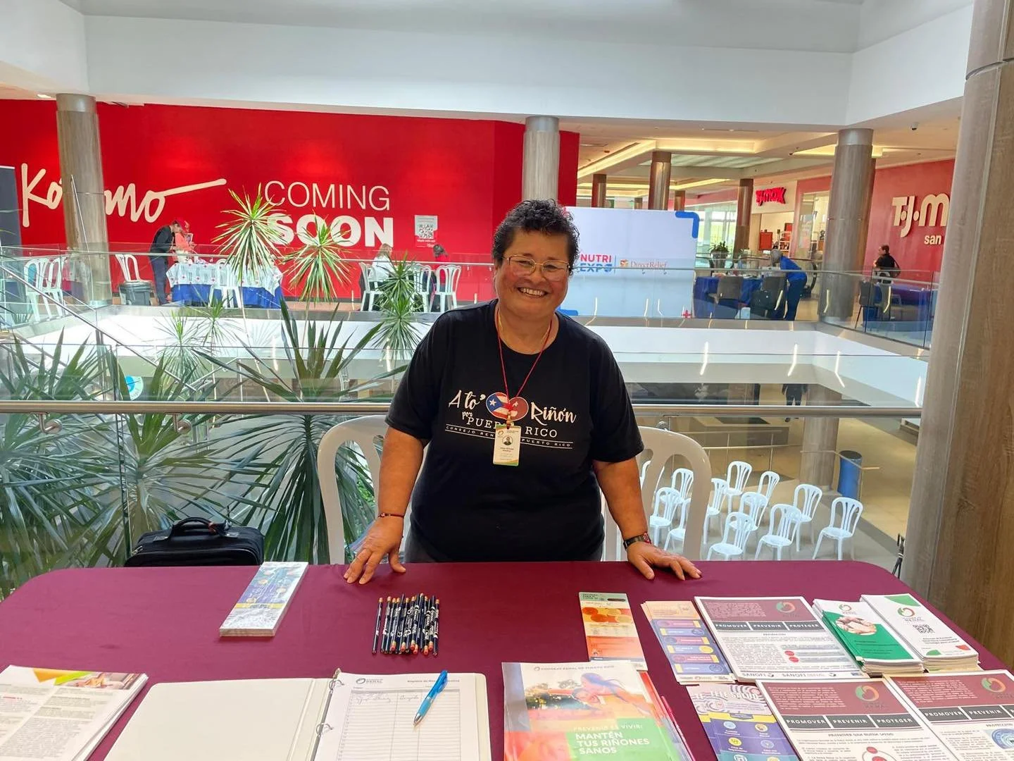 A woman with short curly hair and glasses smiling at a table with pamphlets, pens, and a notebook inside a shopping mall. She wears a black T-shirt with Puerto Rico and a heart symbol. The mall has a red wall with the words 'Coming Soon' and a second