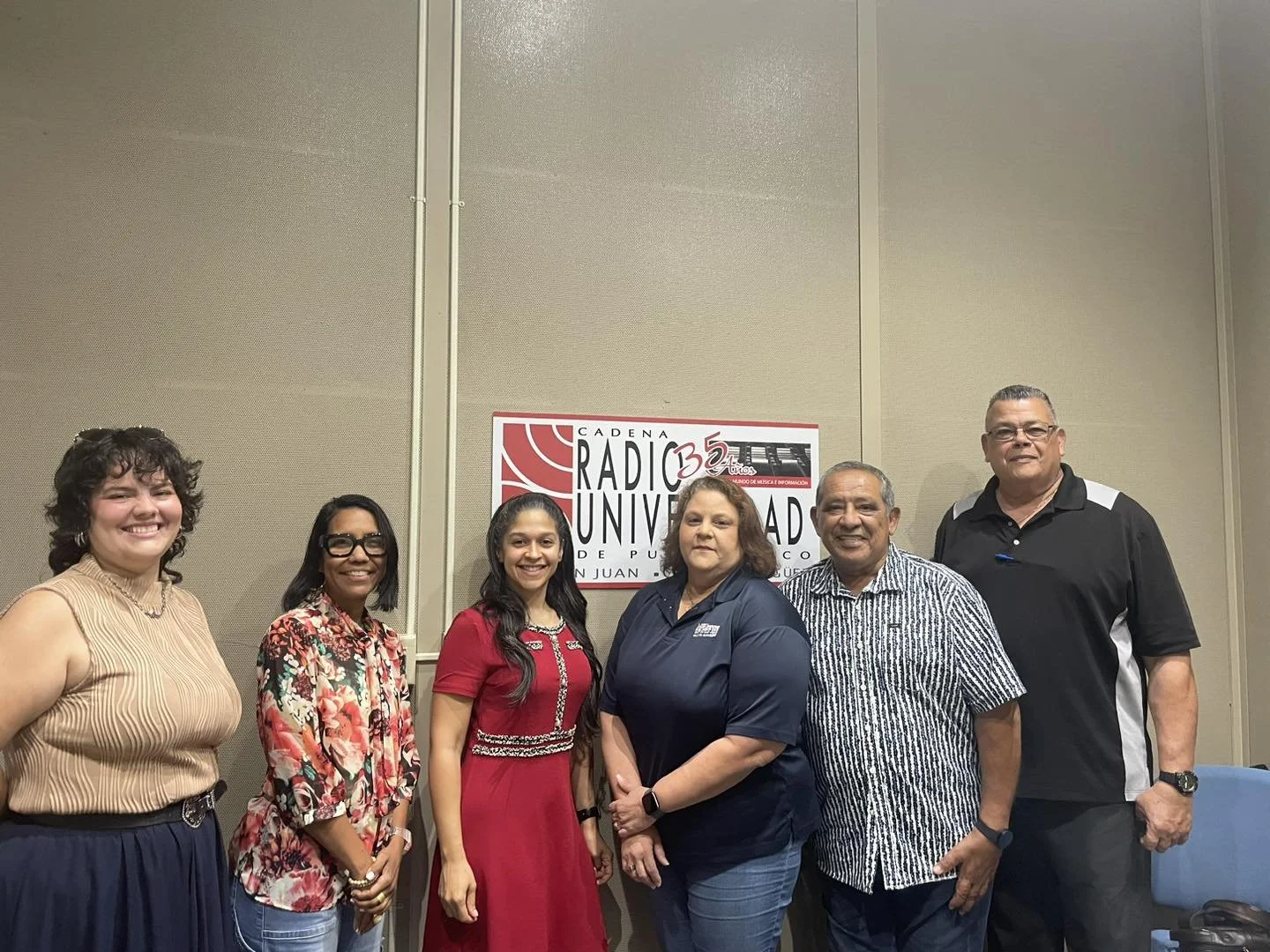 Group of six diverse people standing in front of a sign that reads 'Radio Universidad 35 Aniversario, Cadena Radio Universidad de Puerto Rico, San Juan'.
