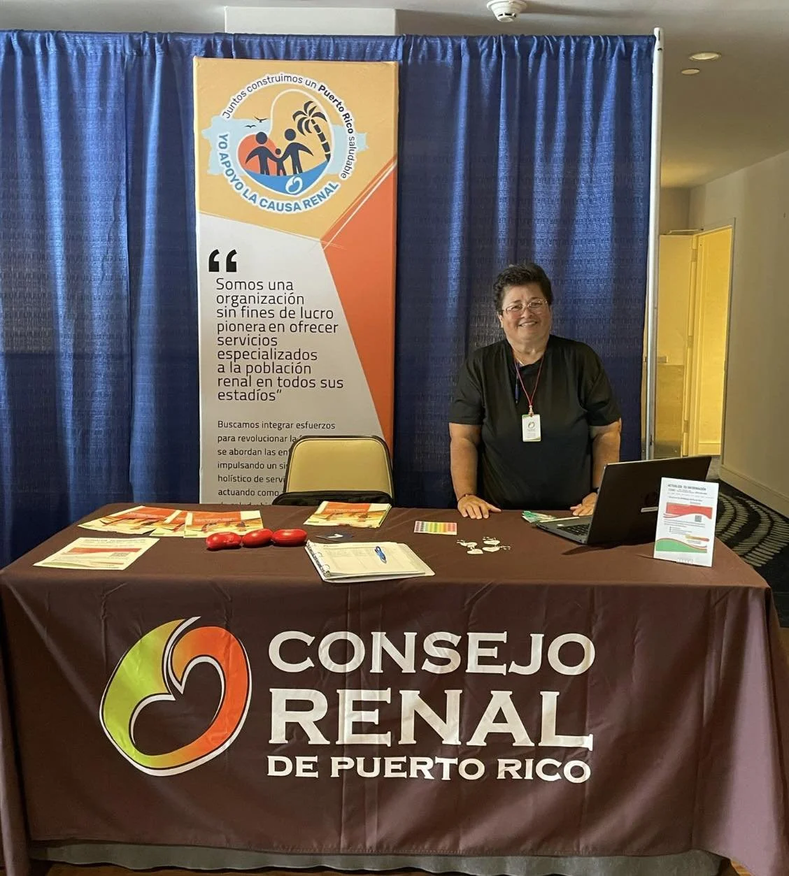 A woman standing at a booth for Consejo Renal de Puerto Rico, with a table covered in a brown tablecloth displaying the organization’s logo and name, and a blue curtain backdrop with a banner explaining their mission to provide specialized renal care