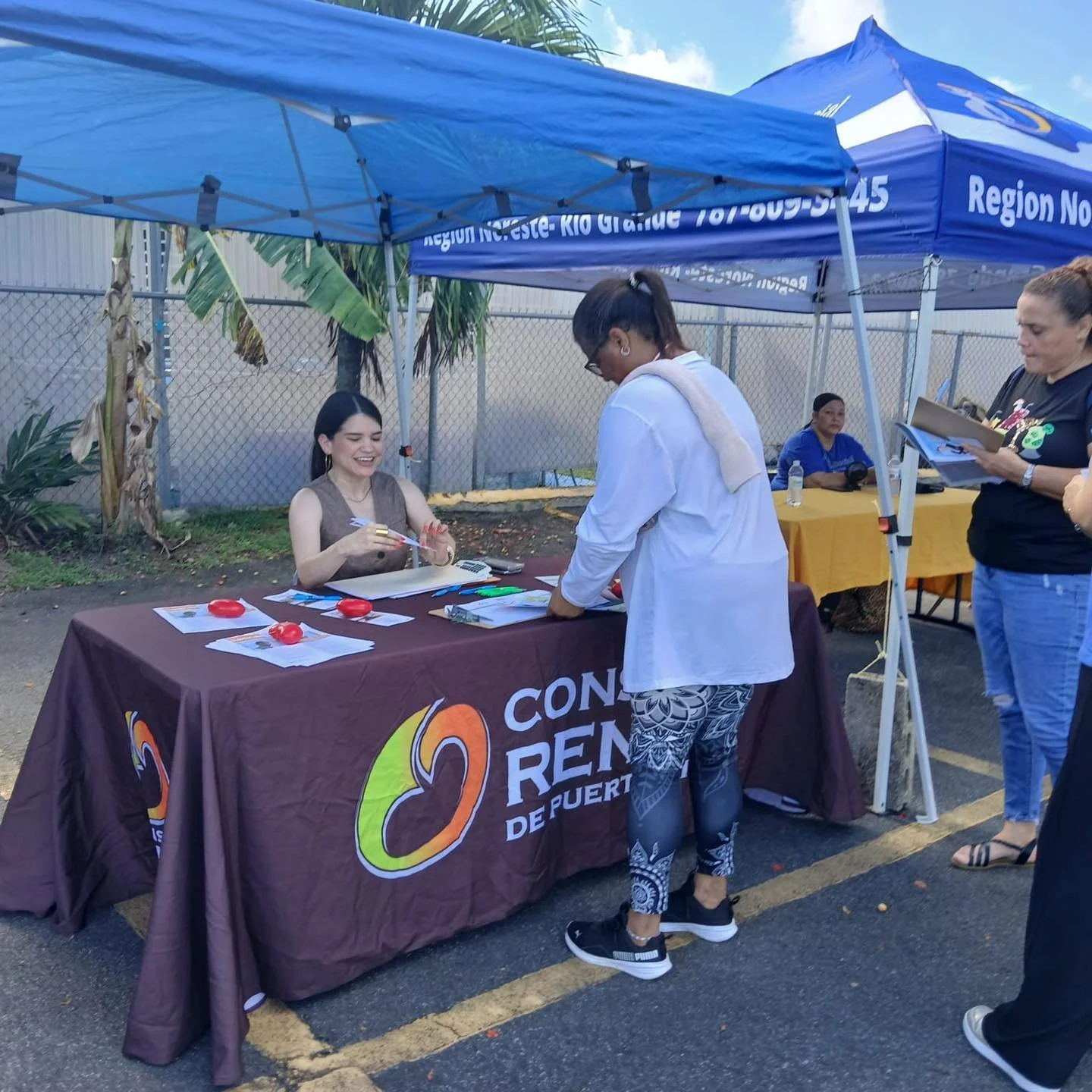 A woman sitting at a table with a dark tablecloth that says "CON RE" and some colorful logo, engaging with a standing woman outside under a blue canopy. There are papers and red objects on the table. Several other people are nearby, some taking notes
