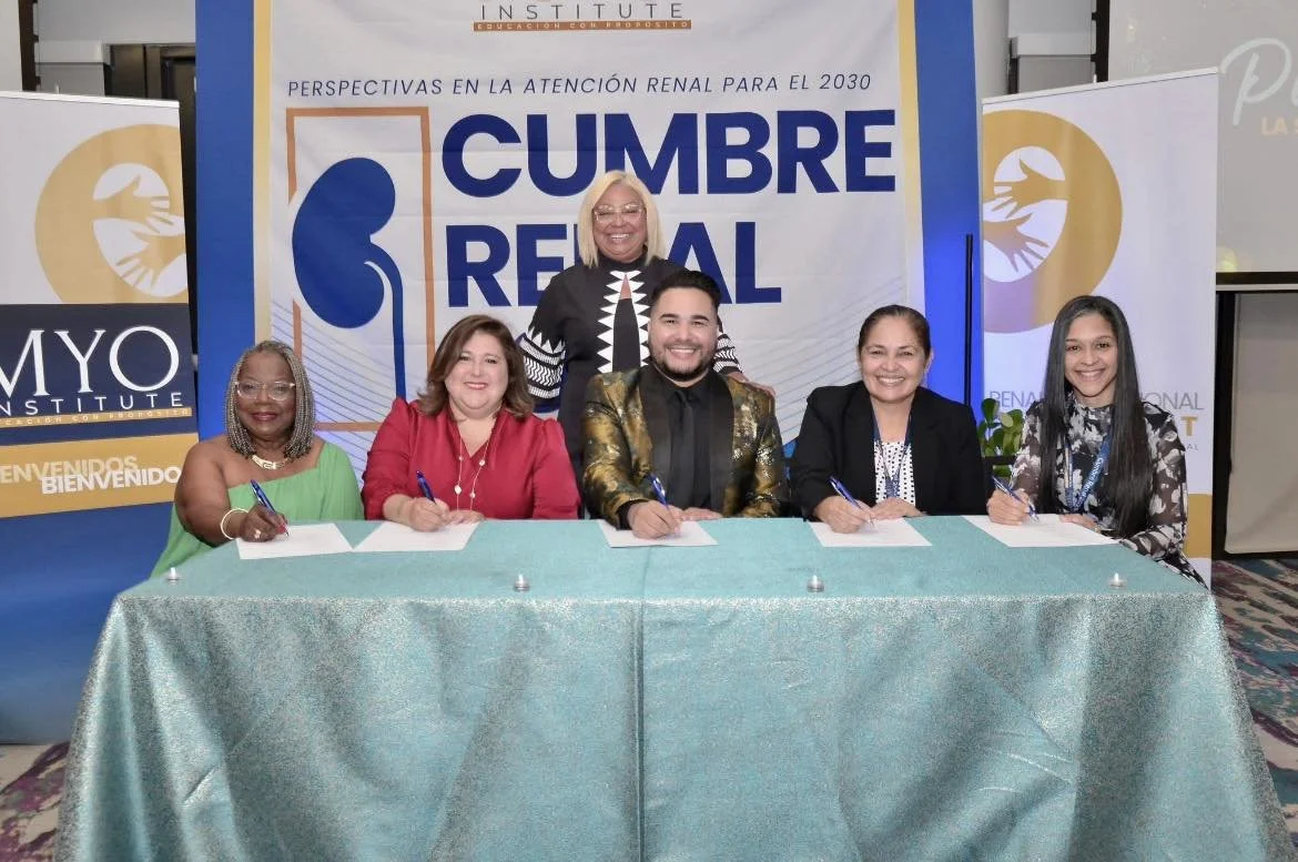 Six diverse people sitting at a table signing documents at a conference event with banners and a large backdrop behind them.