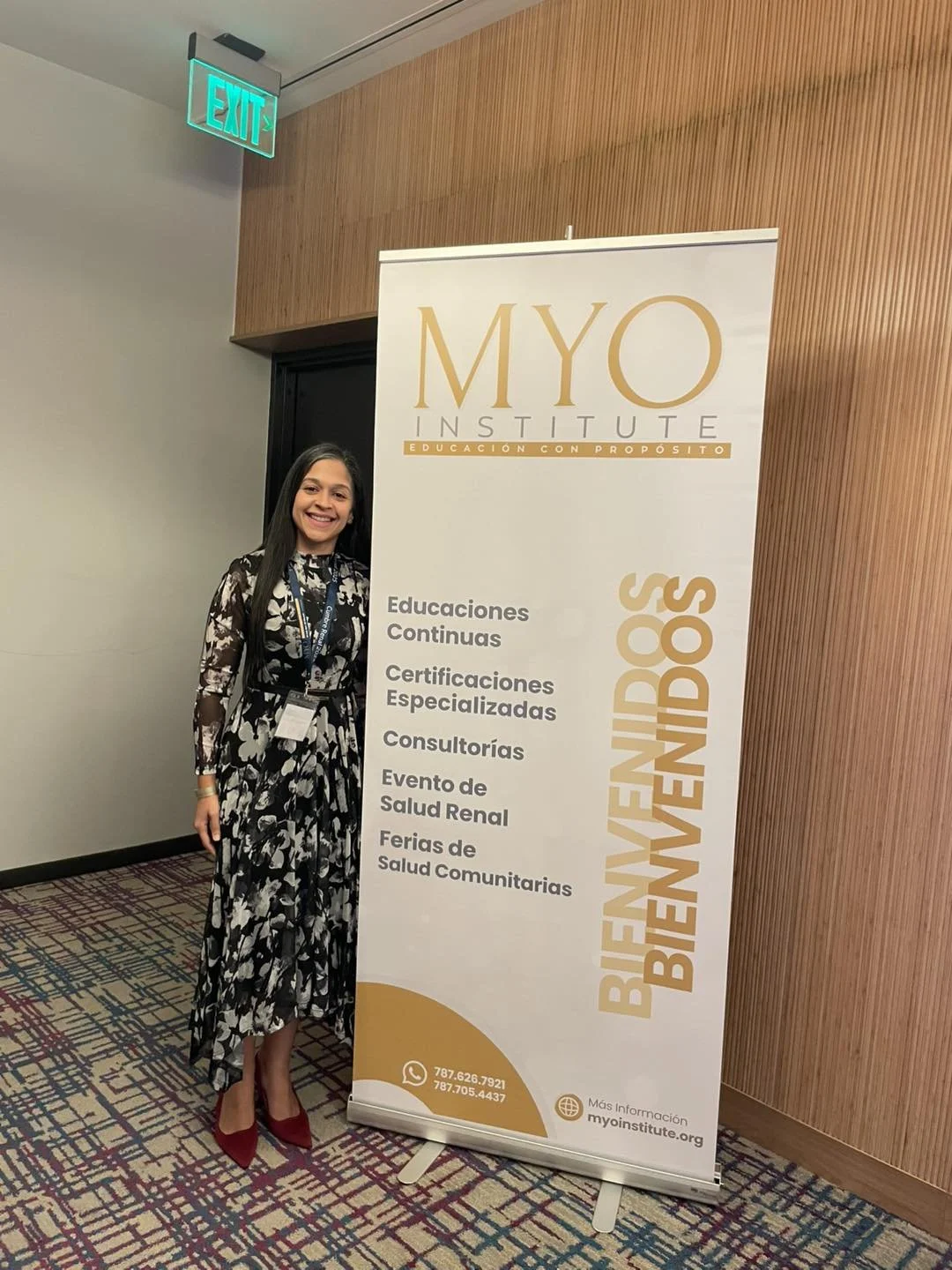 A woman standing next to a large white vertical banner at an indoor event, smiling. She is wearing a black and white floral dress, red high heels, and a lanyard with a badge. The banner displays information about MYO Institute, including services lik