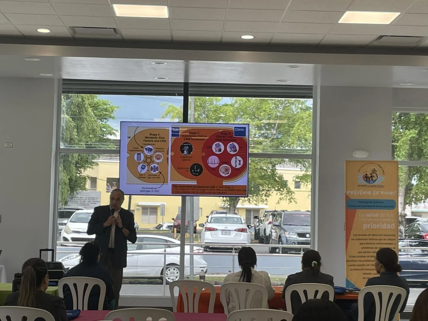 A man in a suit giving a presentation in a conference room with a large screen displaying a medical infographic. Several women in business attire sit at colored tables, facing the speaker. Large windows overlook parked cars and trees outside.