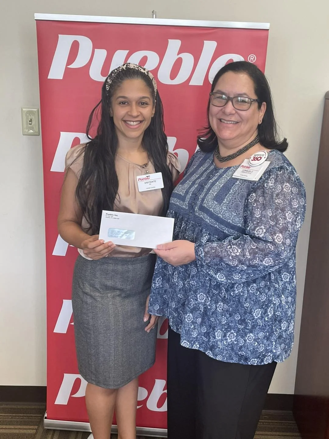 Two women standing in front of a pink Pueblo banner, smiling and holding an envelope, at an event or awards ceremony.