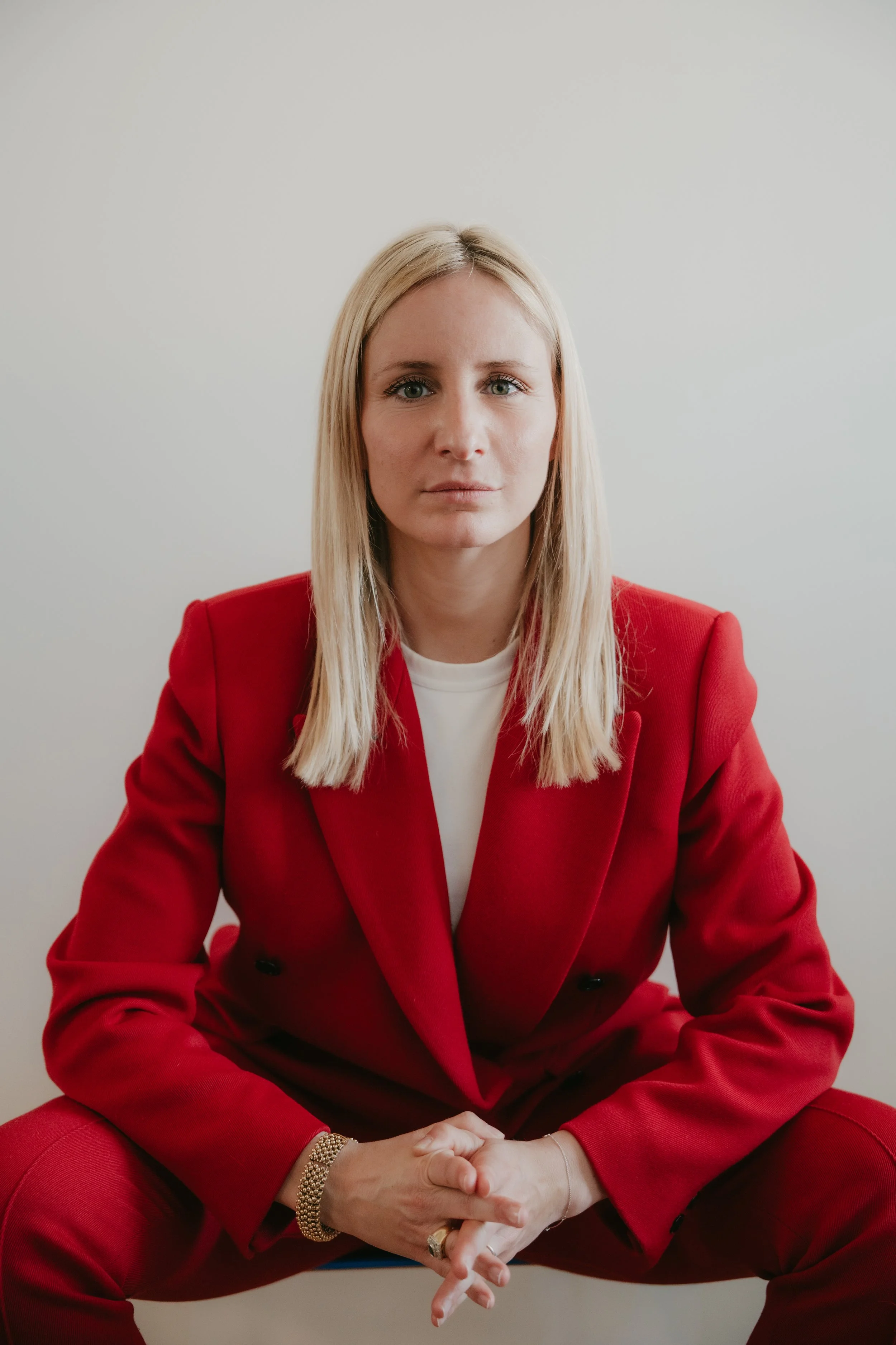 Femme blonde assise, portant un tailleur rouge, devant un mur blanc.