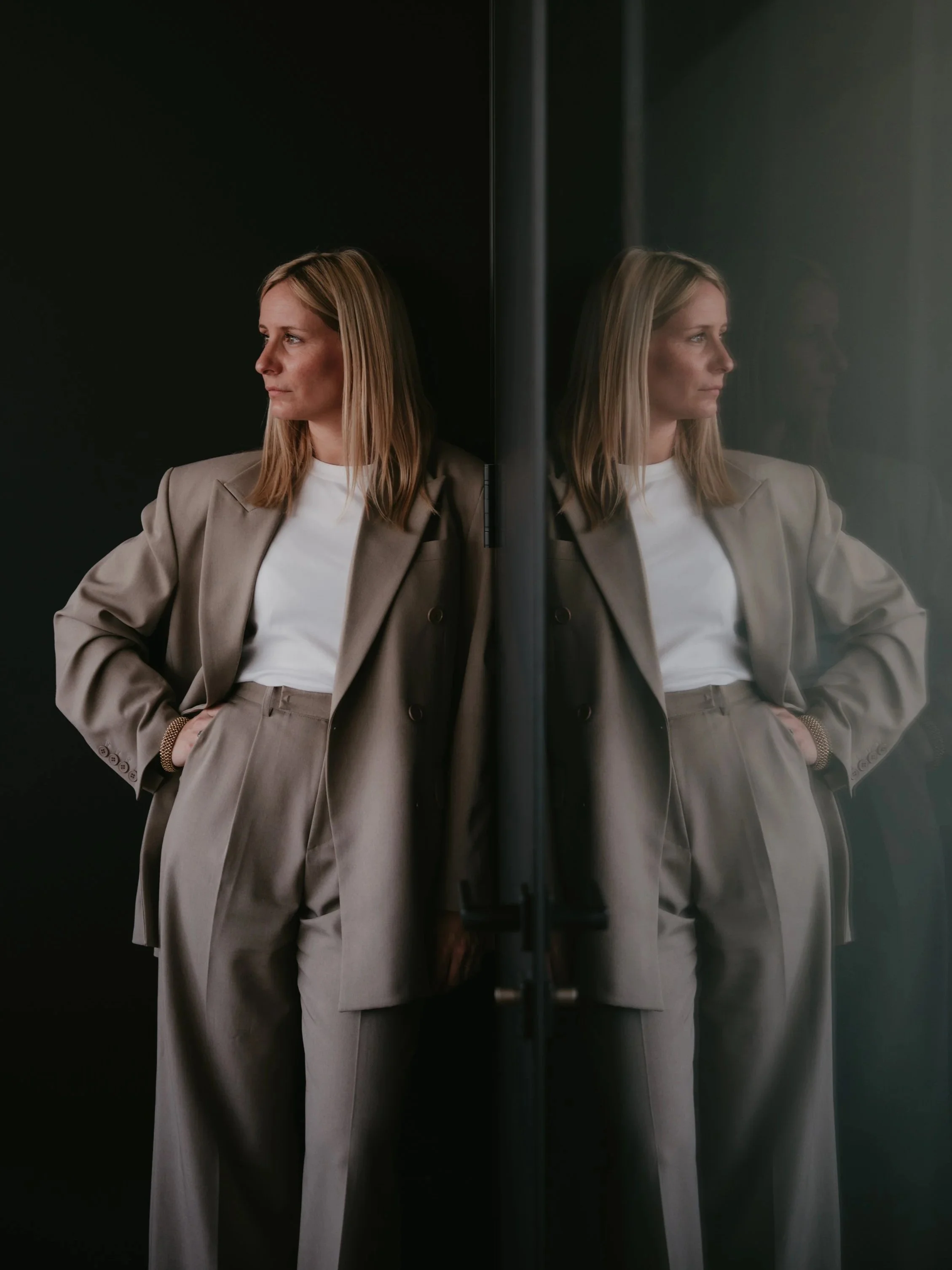 Une femme blonde portant un tailleur gris clair et un t-shirt blanc, debout devant une vitre, vue de profil, regardant vers la gauche.