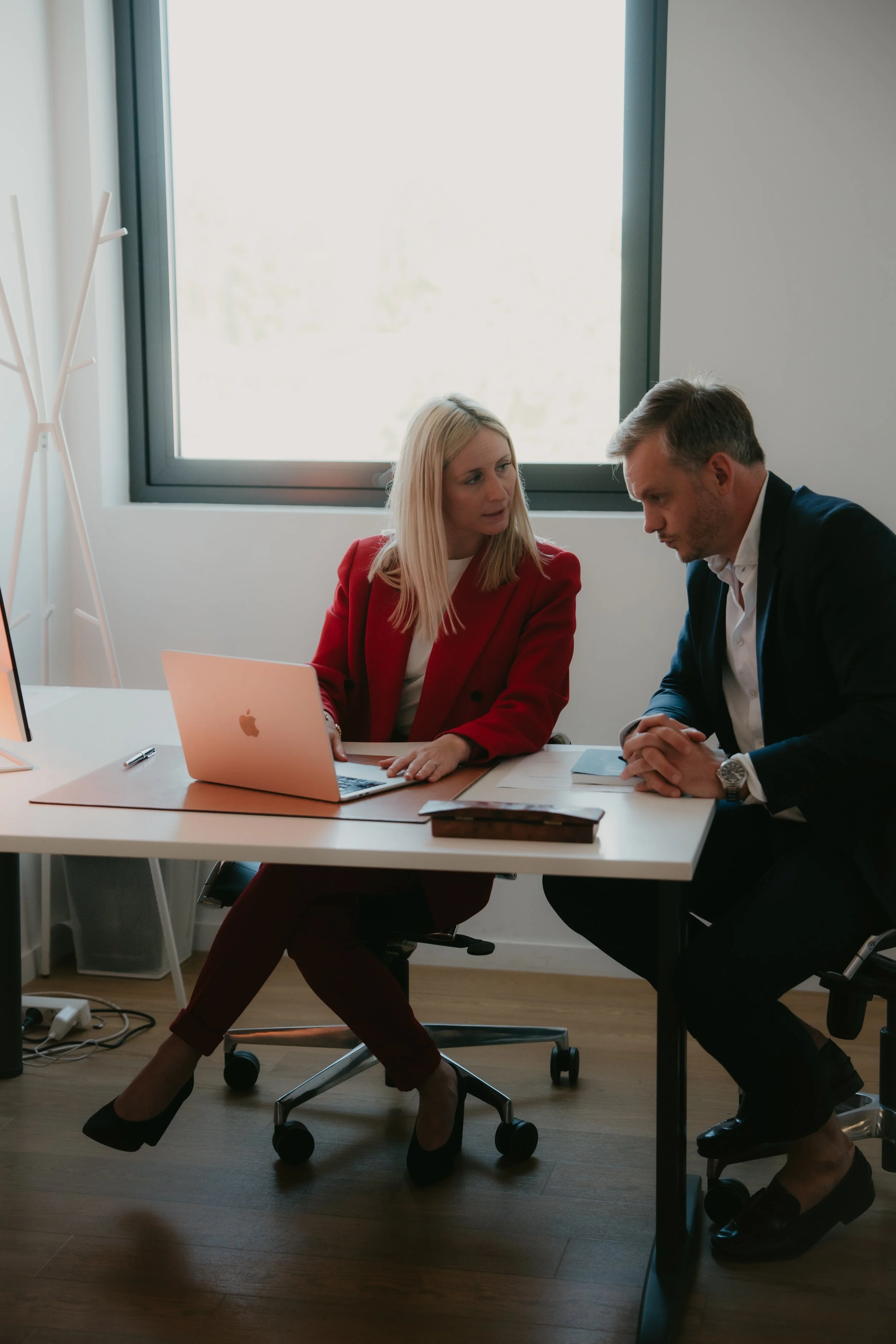 Deux professionnels discutent dans un bureau moderne. La femme porte une veste rouge et des talons noirs, assise devant un ordinateur portable rose_OR.