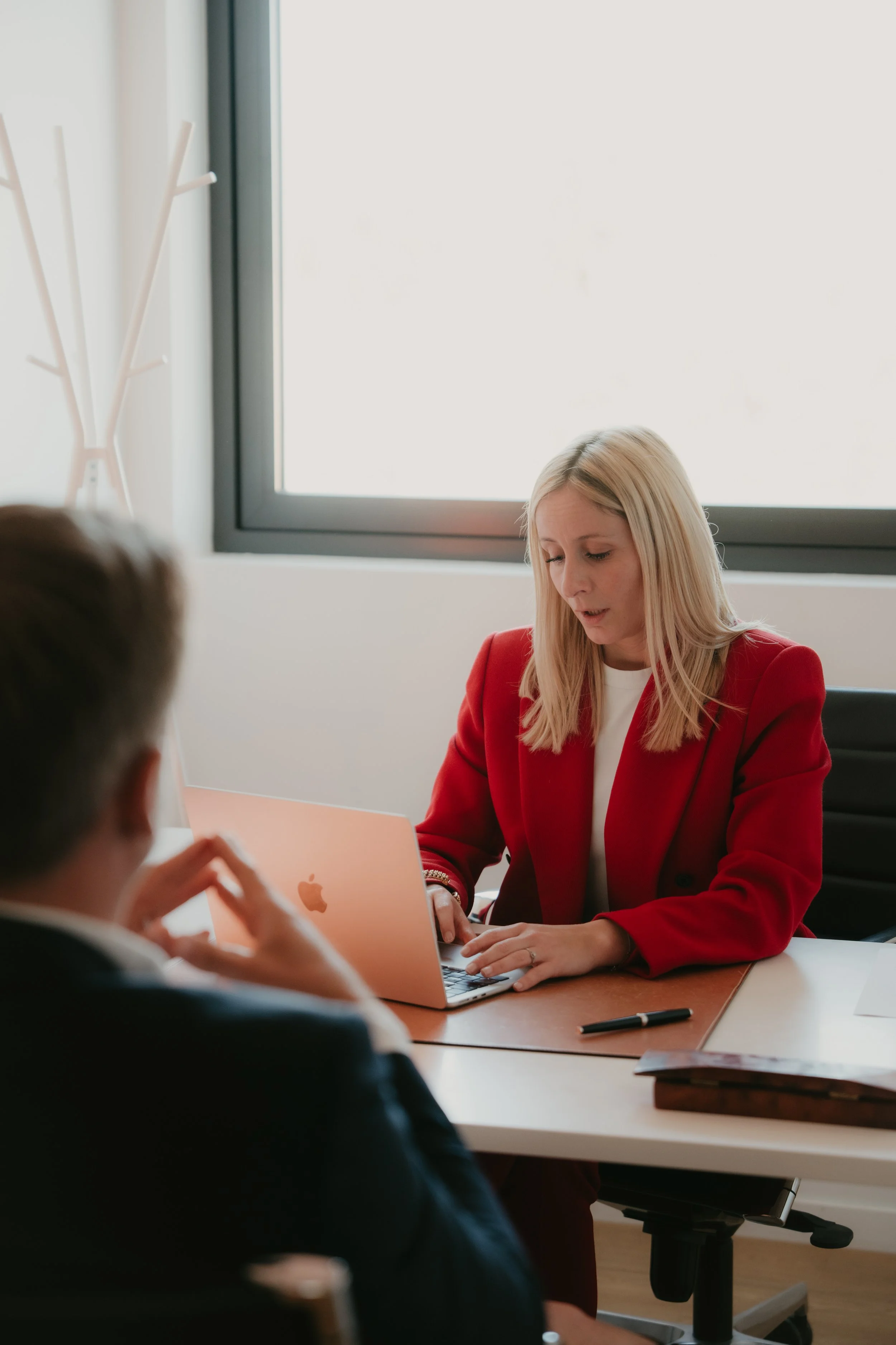 Une femme blonde en blazer rouge travaillant sur un ordinateur portable lors d'une réunion, discutant avec deux collègues dans une salle de réunion moderne.