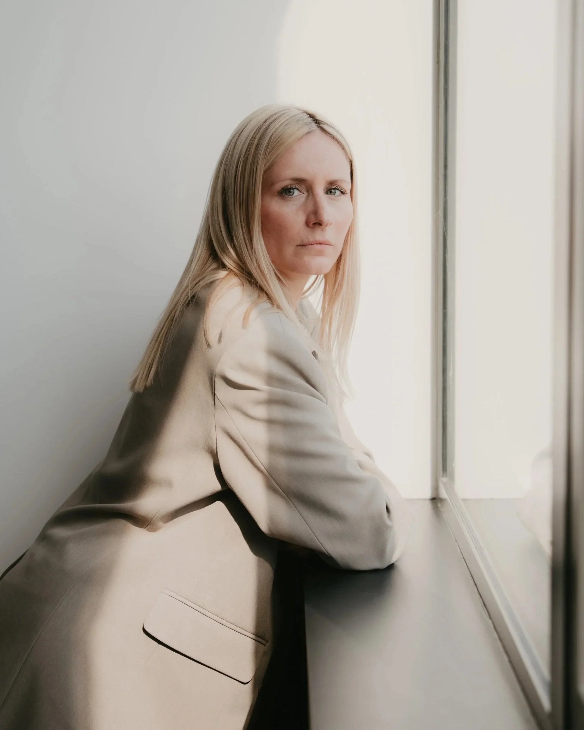 Femme aux cheveux blonds, portant une tenue beige, assise sur le sol près d'une fenêtre dans un espace moderne, regarde vers l'objectif.