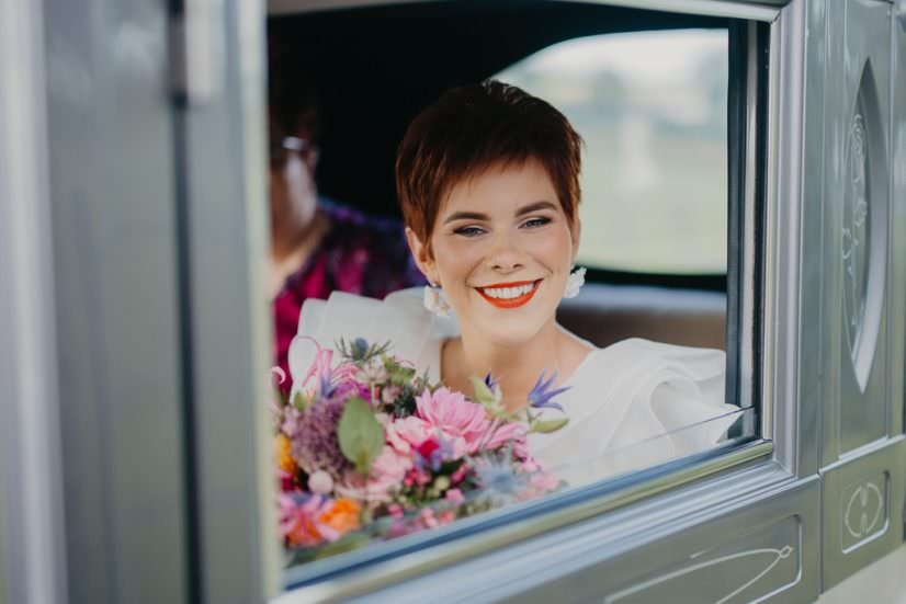 A woman with short brown hair, wearing white and bright red lipstick, smiling through a bus window while holding a colorful bouquet of flowers.