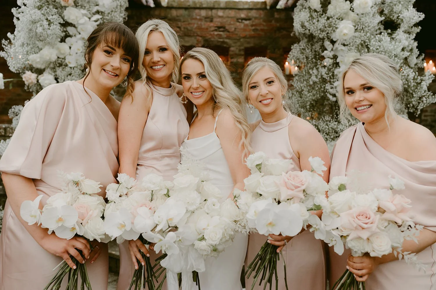 Group of five women at a wedding reception, dressed in light pink and white attire, holding bouquets of white and blush roses and orchids, standing in front of a decorative floral backdrop.