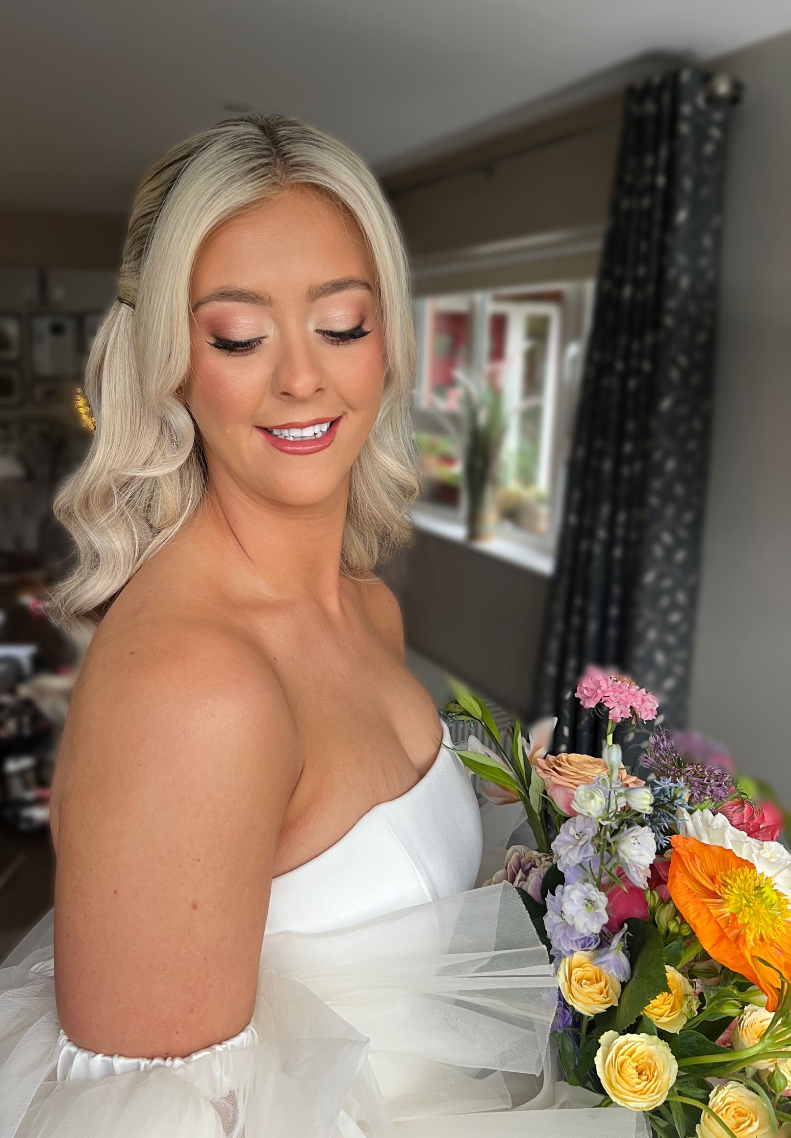 A woman with blonde hair smiling, wearing a white lace dress with floral patterns, standing indoors with a blurred background.