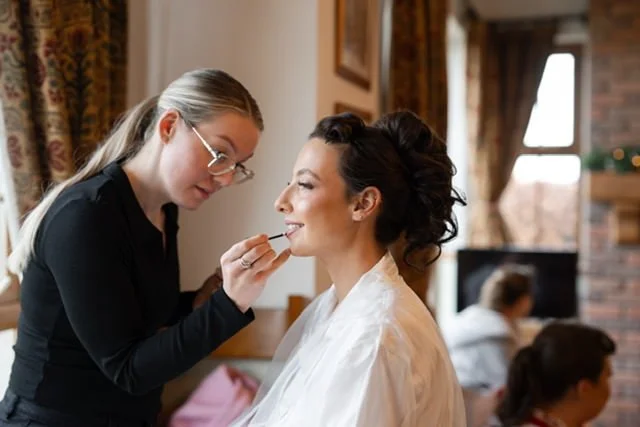 A makeup artist applying makeup to a woman's face in a cozy, well-lit room.