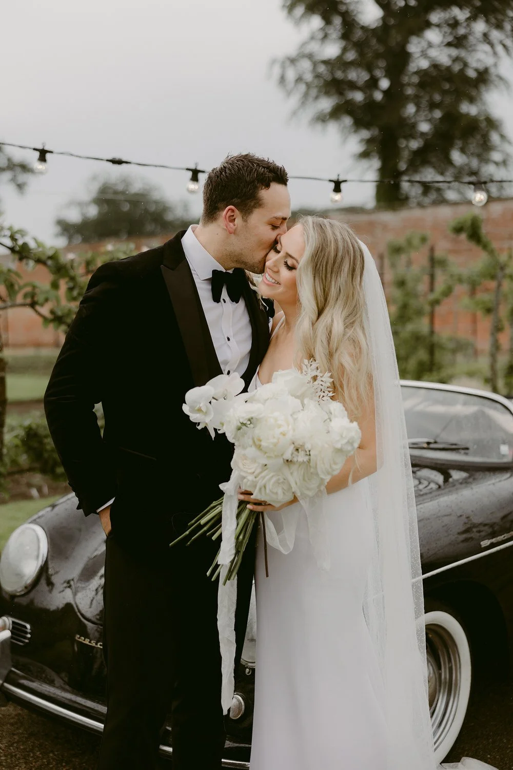 Bride and groom sharing a kiss, with the groom dressed in a black tuxedo and the bride in a white wedding gown holding a bouquet of white flowers, standing outdoors near a vintage black car and string lights.