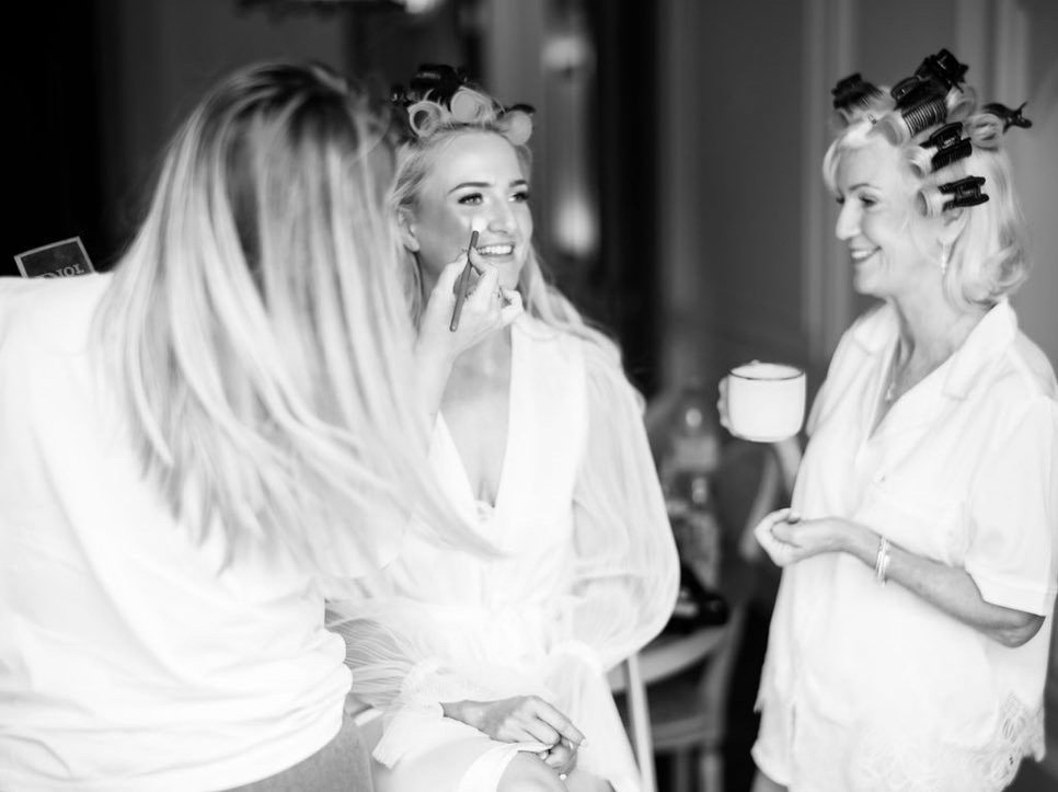 Women with hair rollers getting makeup applied, smiling, in a room with a makeup artist holding a brush.