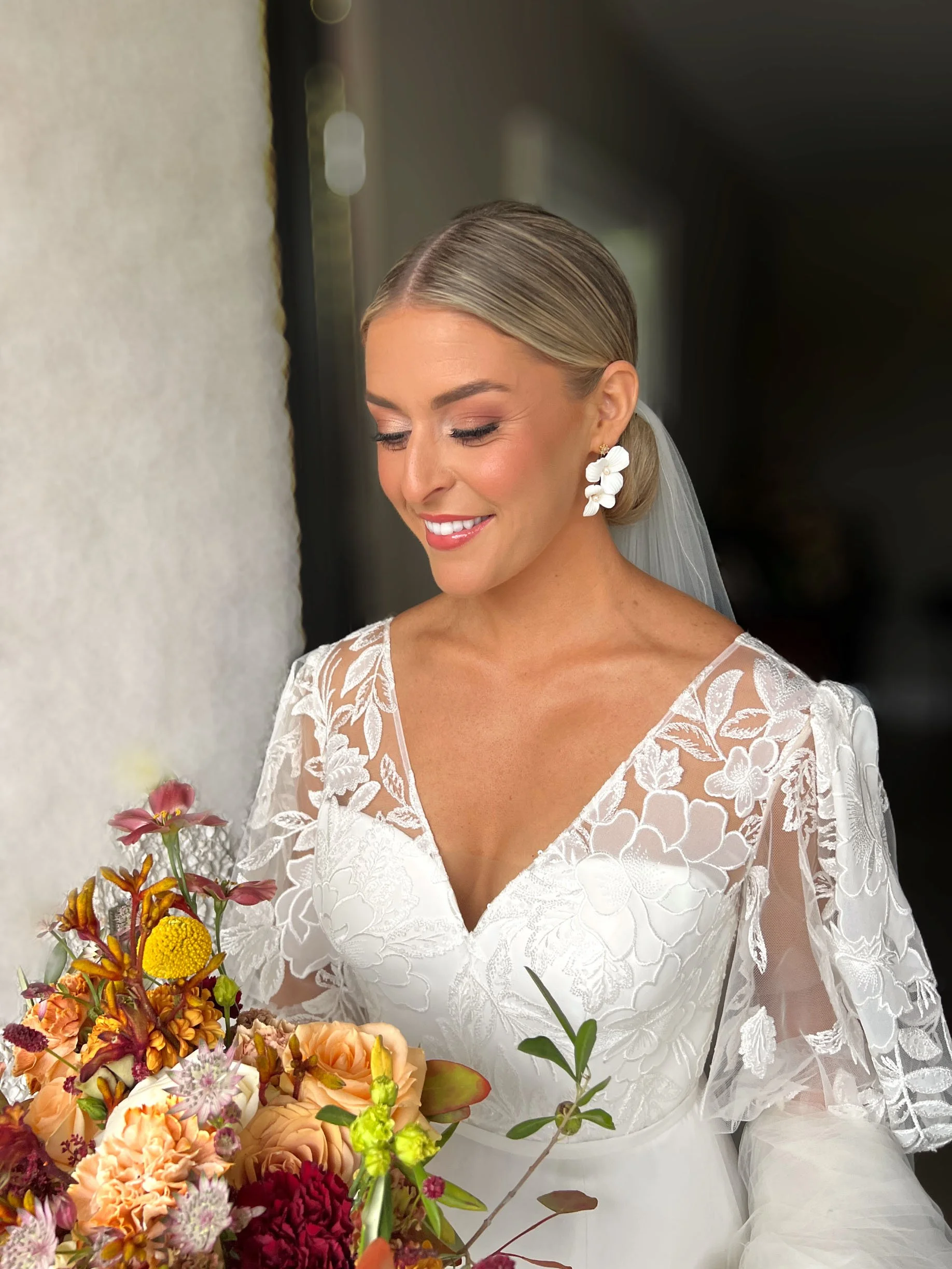 A smiling bride holding a bouquet of pink, white, and purple flowers, wearing a white off-shoulder wedding dress and veil.